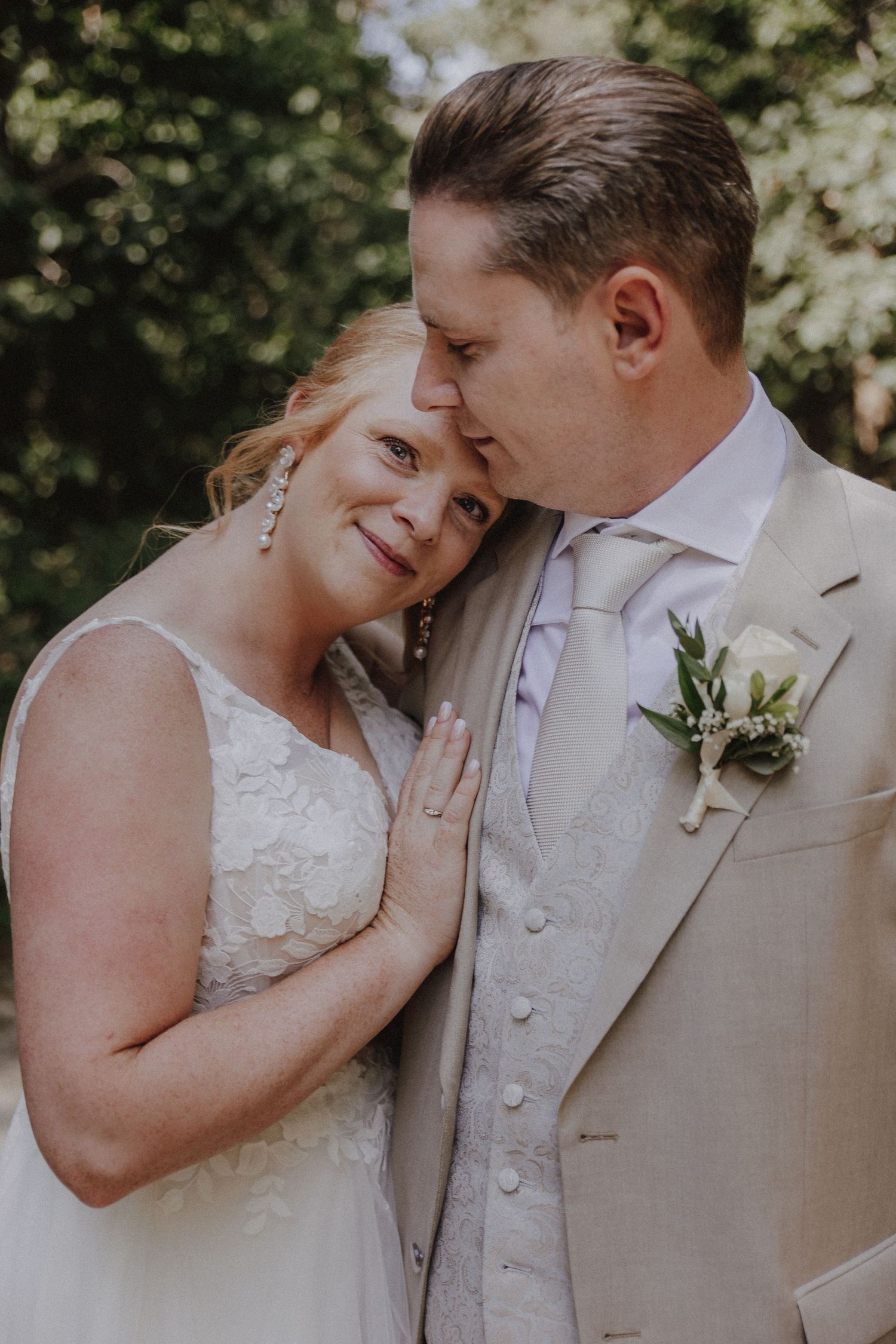 Bride in lace gown leans on groom in cream suit with boutonniere during romantic outdoor wedding portrait.