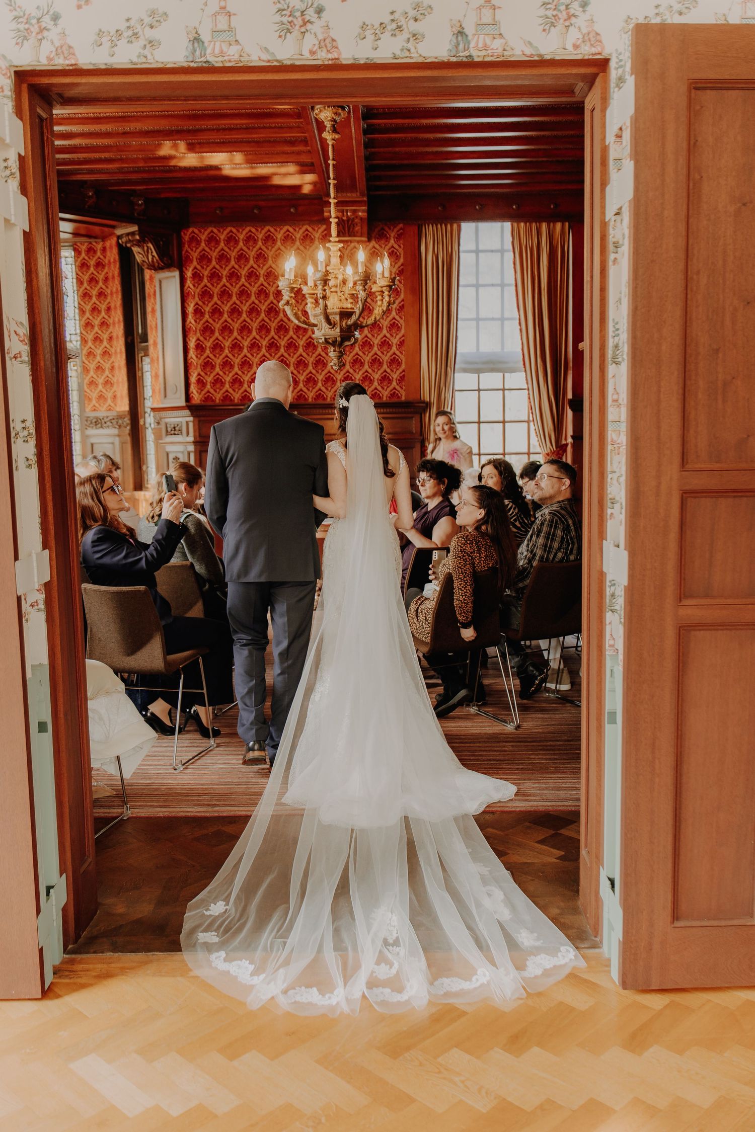 Bride in white gown with long veil stands at doorway of elegant wedding ceremony room with chandelier.