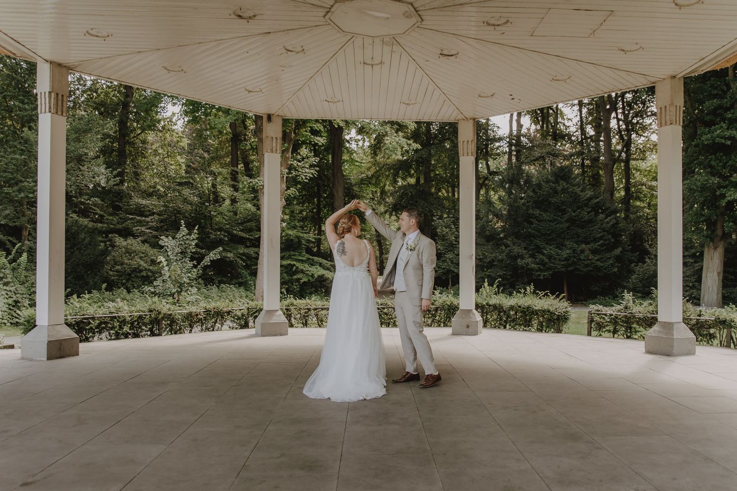 Bride and groom sharing a romantic moment under a white open-air pavilion surrounded by lush trees.