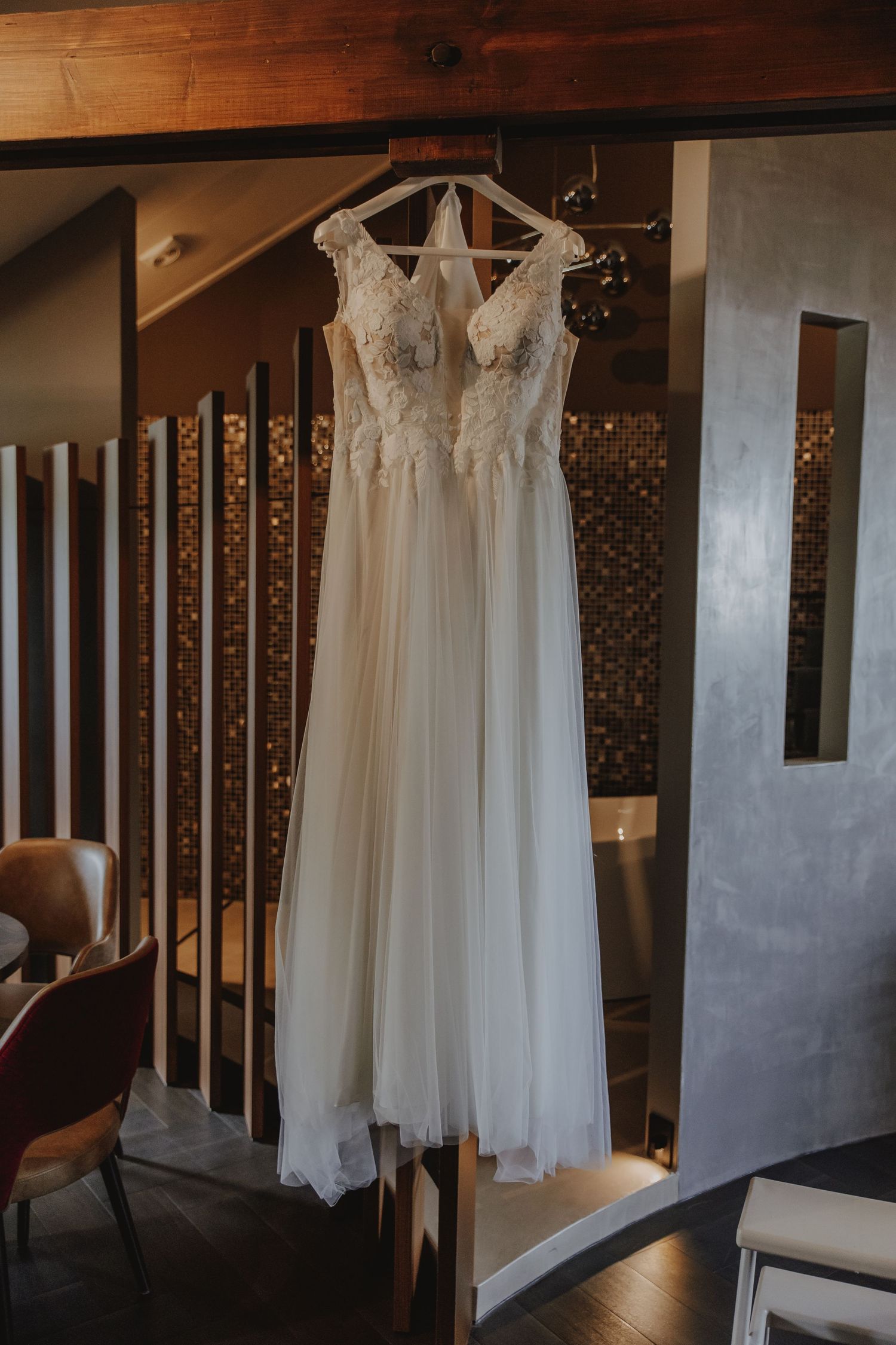 Elegant lace bodice wedding dress hanging on a wooden hanger in a warmly lit rustic interior.