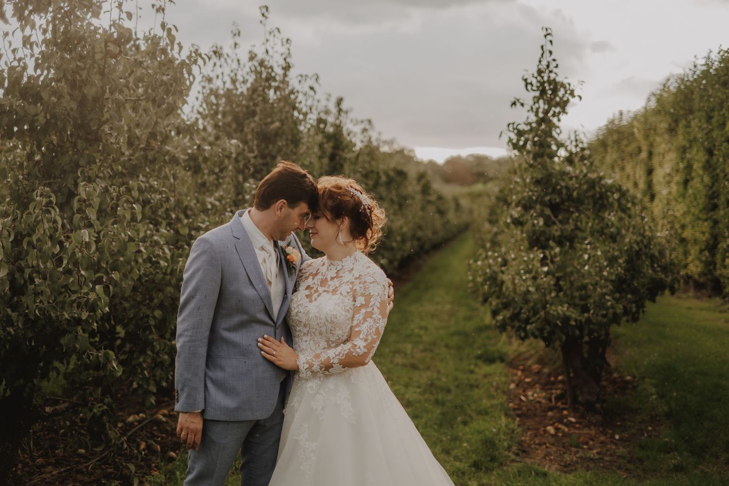 Bride and groom share romantic moment in lush apple orchard, wearing elegant wedding attire on a cloudy autumn day.
