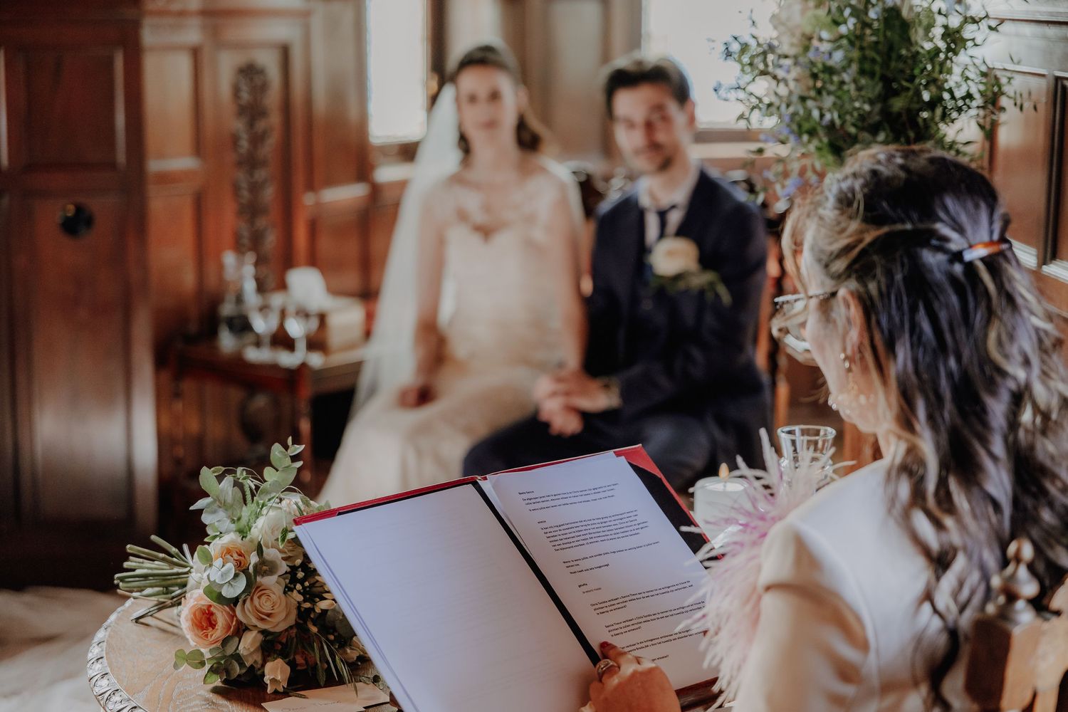 Officiant reads from red binder during intimate wedding ceremony with bride and groom in background.