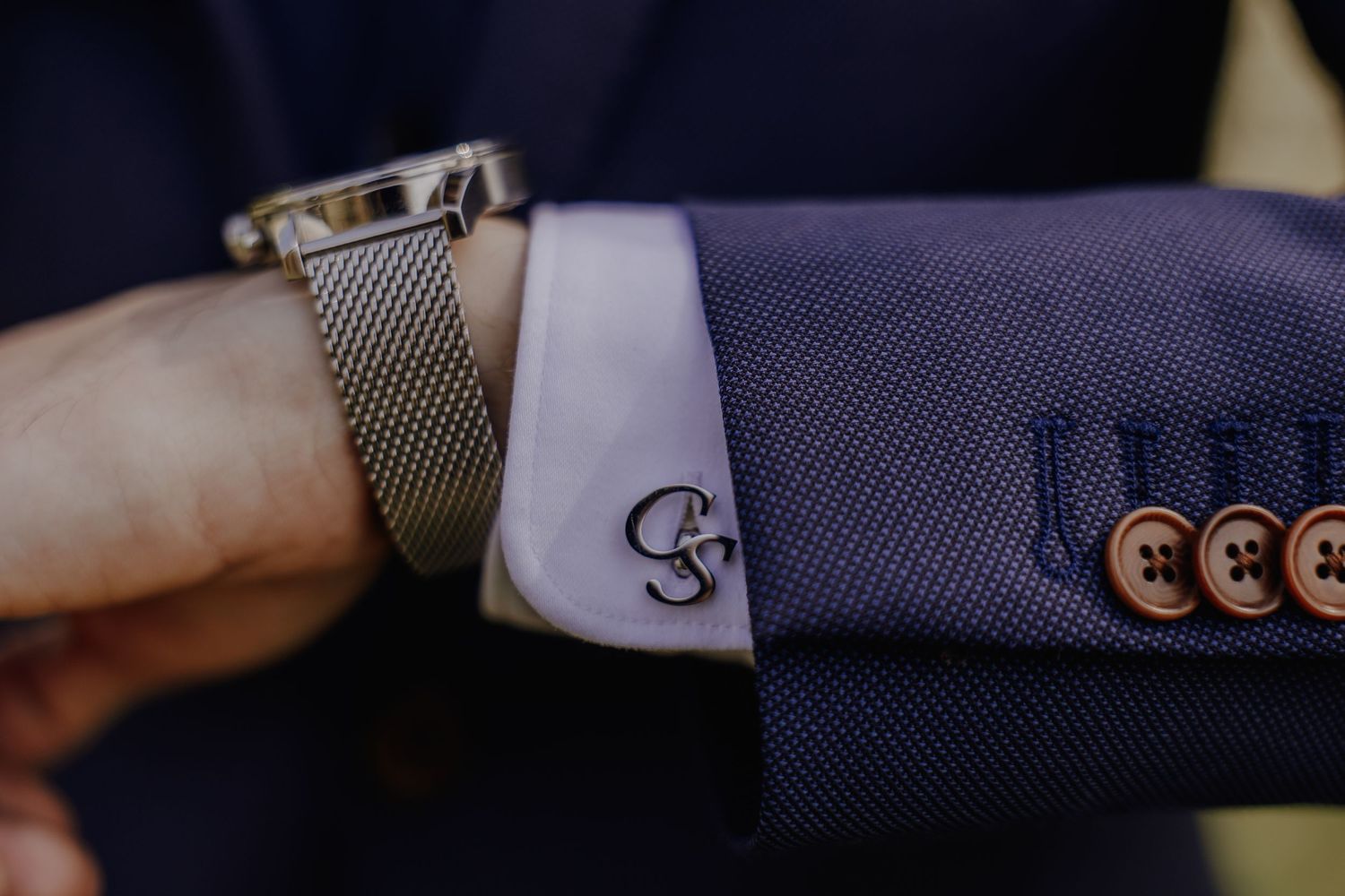 Close-up of a man's wrist wearing a silver mesh watch and monogram cufflink on a navy blue suit jacket.