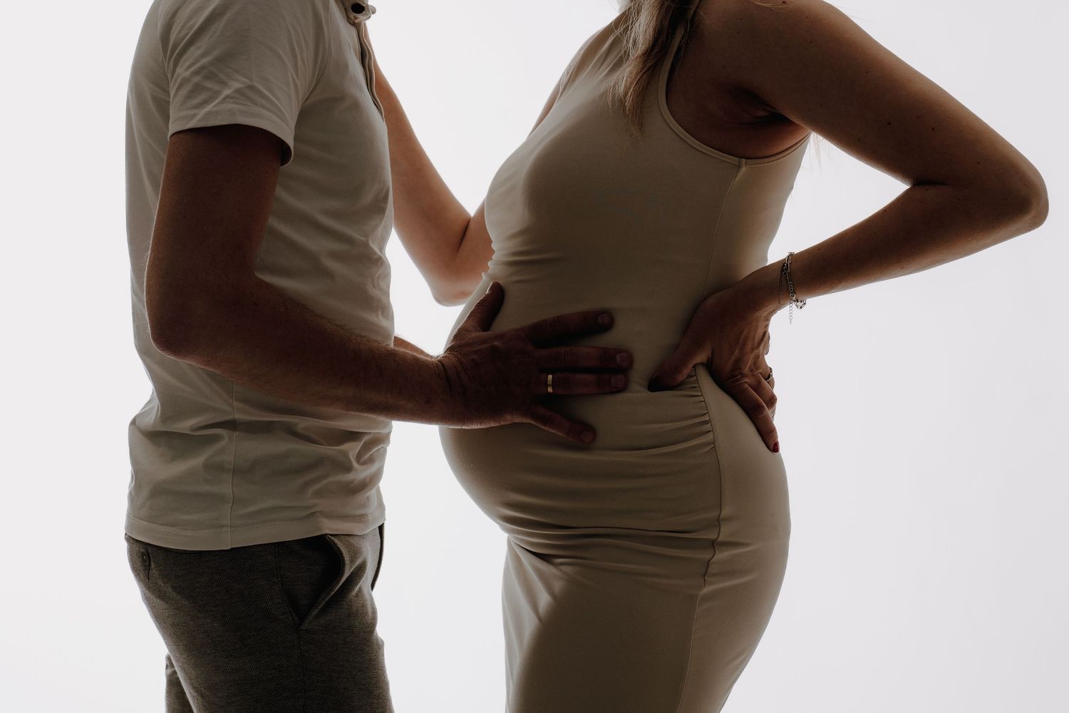 A pregnant woman in a beige dress stands with her partner, both cradling her baby bump against a light background.