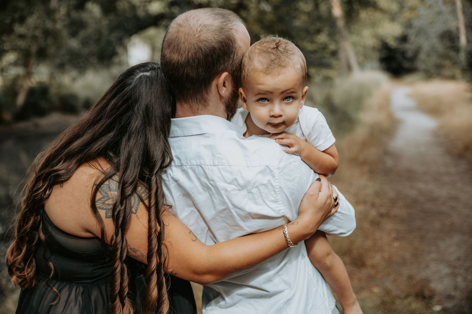 A couple holds their young toddler on a forest path, the child looking back at the camera.