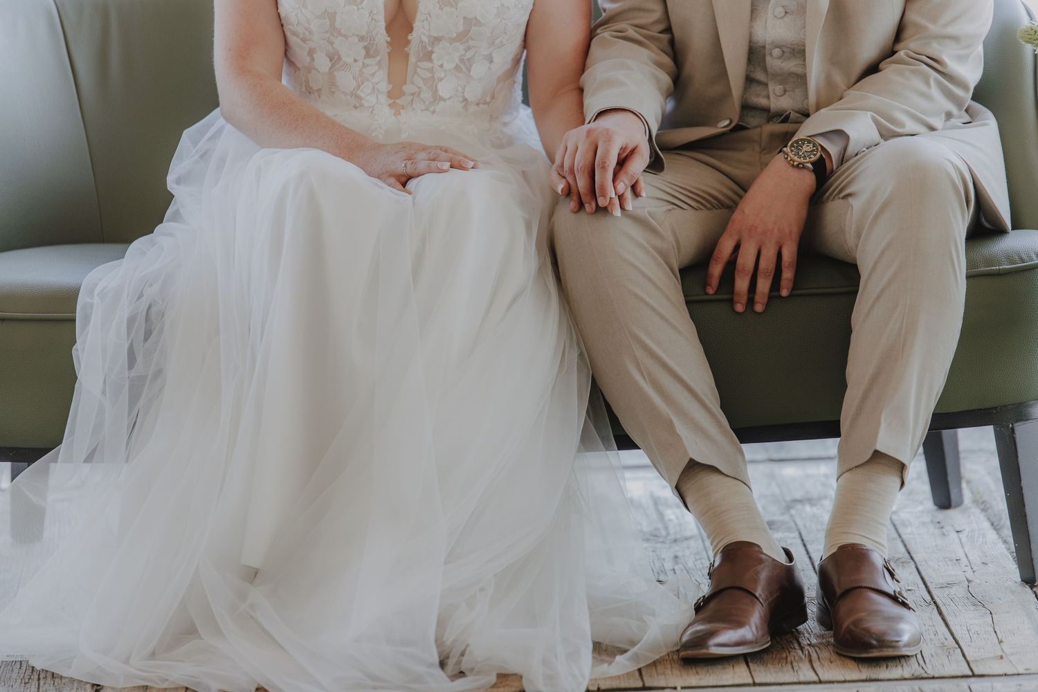 Bride in white tulle gown and groom in tan suit sitting together holding hands on a green velvet sofa.