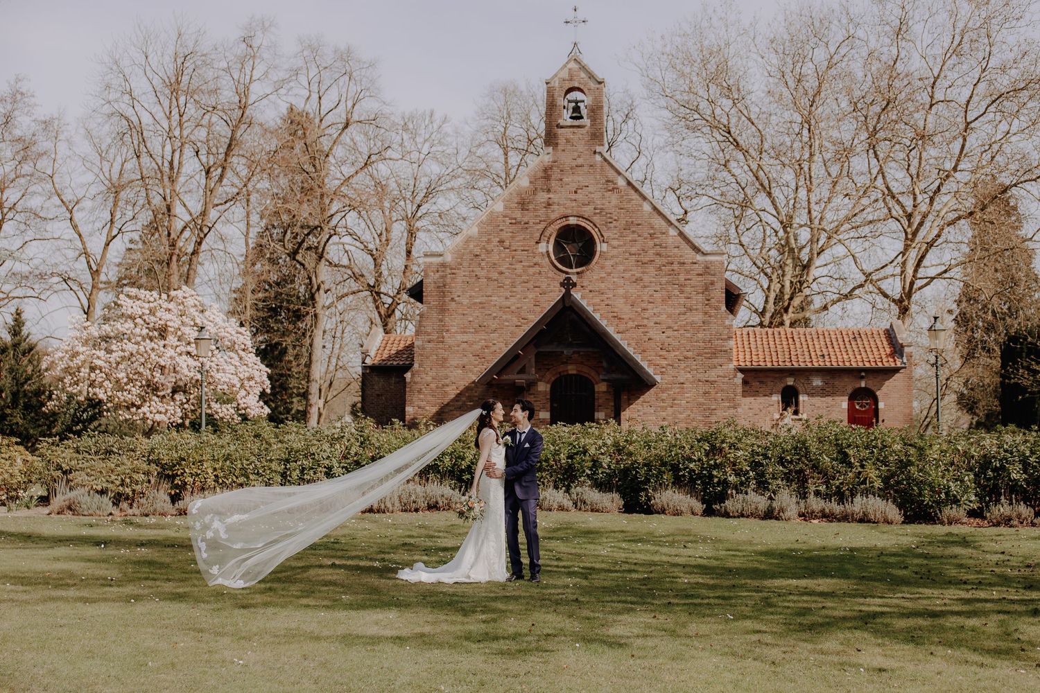 Wedding couple embracing in front of a brick chapel with brides veil flowing in the wind on a spring day.