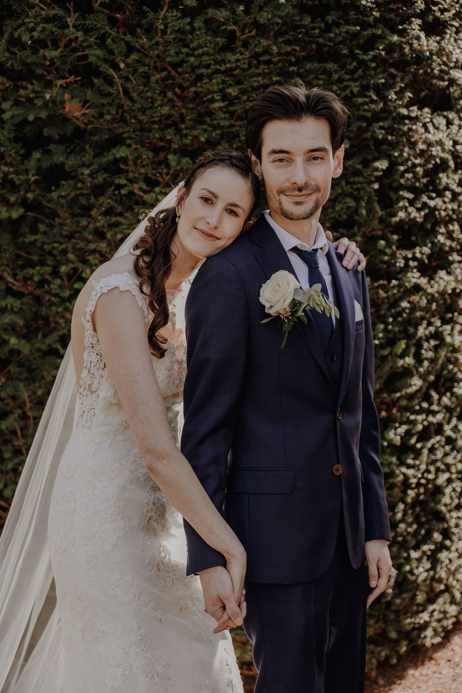 Bride and groom posing together outdoors, holding hands in front of green hedge.