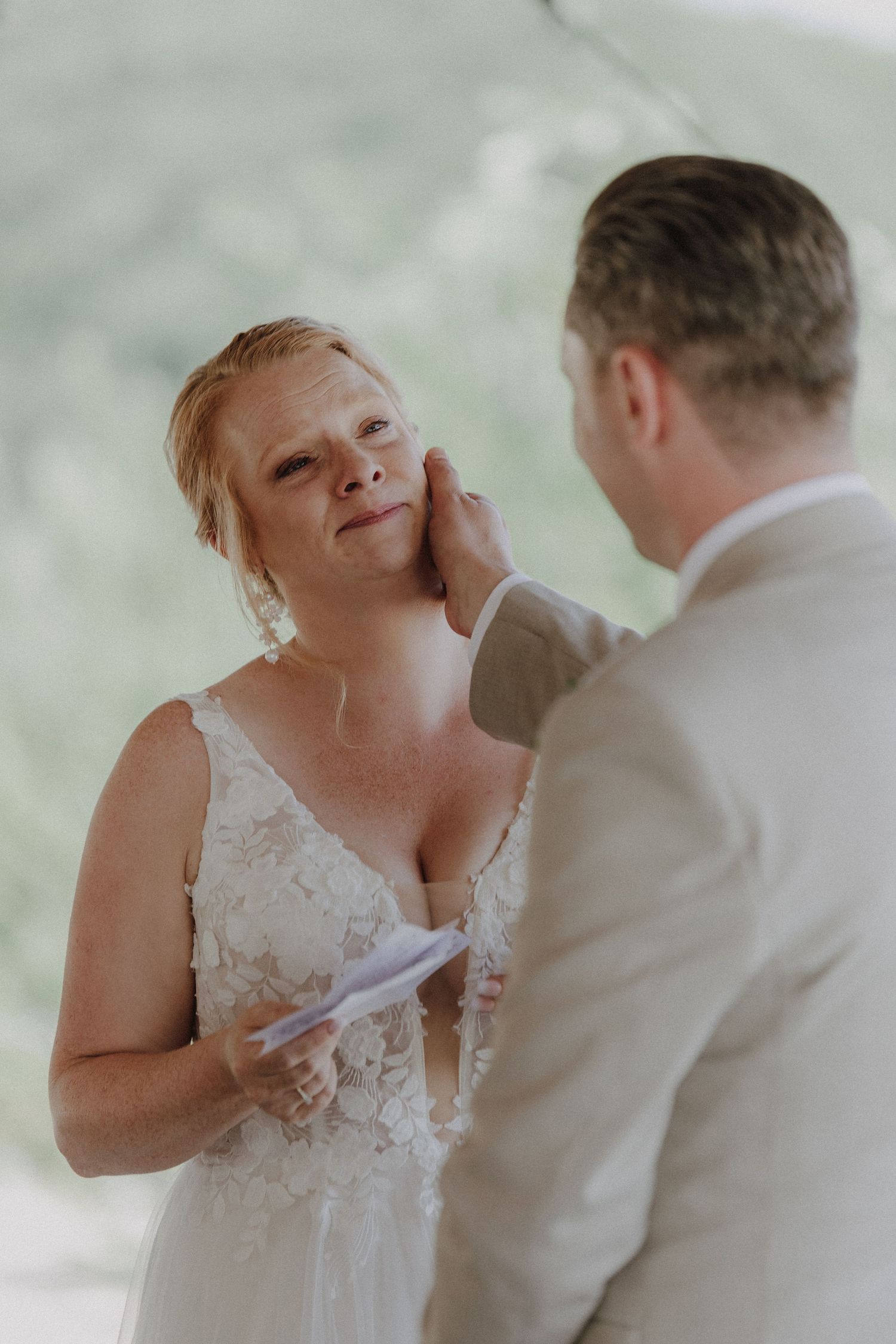 Emotional bride in lace wedding dress reading vows while groom gently touches her face outdoors.