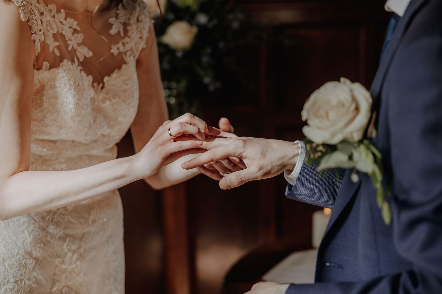 Bride placing a wedding ring on grooms finger during ceremony, close-up of hands with white rose boutonniere.
