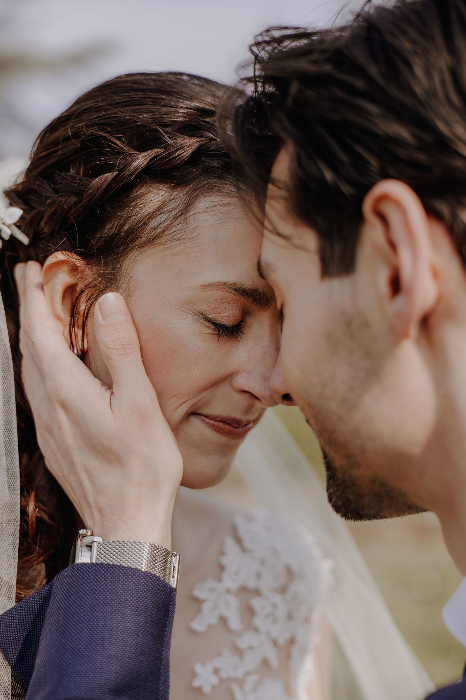 Groom gently cradling brides face as they touch foreheads, bride in lace wedding dress with braided hair.