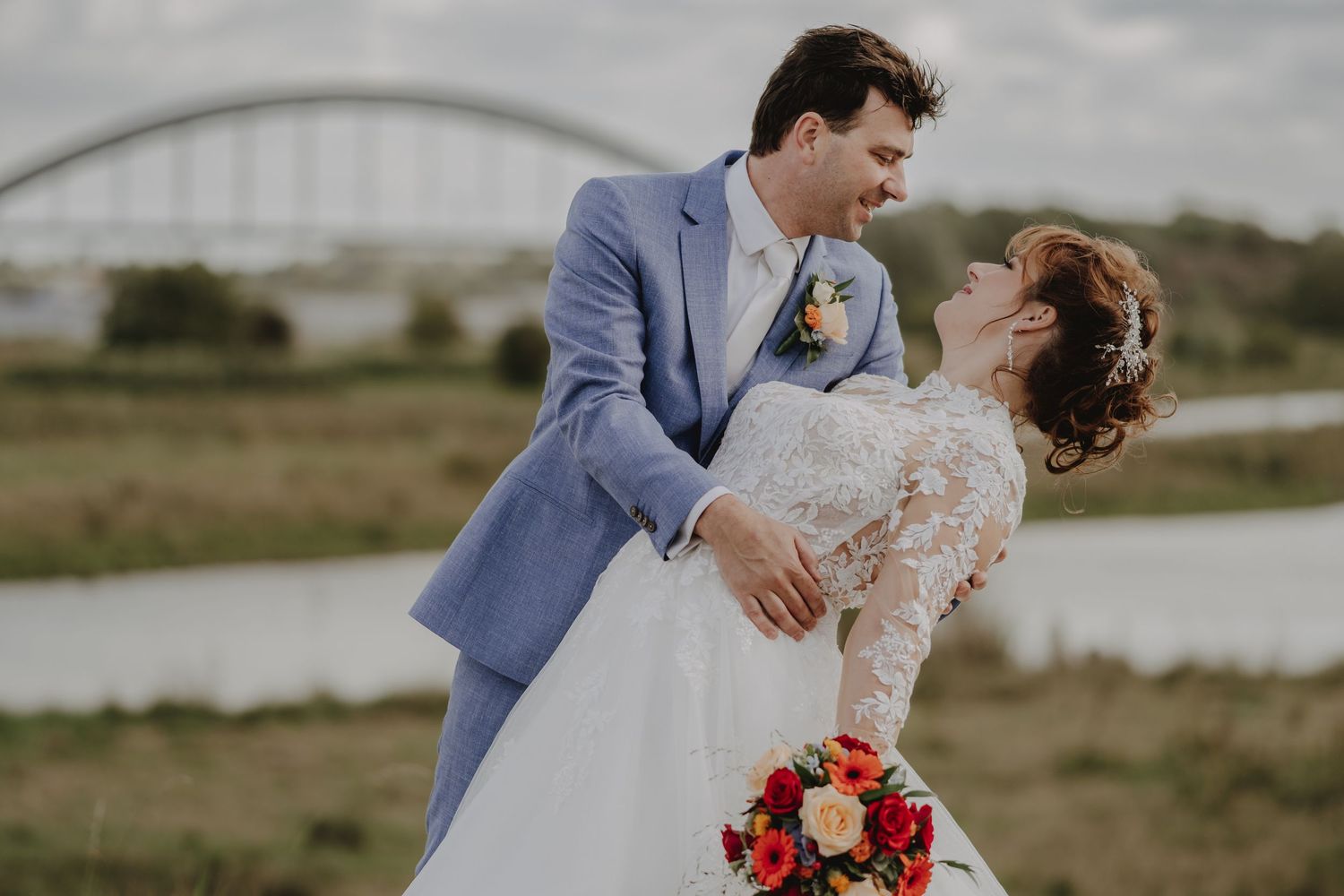 Bride and groom share a romantic dance outdoors near a river bridge, bride holding a vibrant red bouquet.