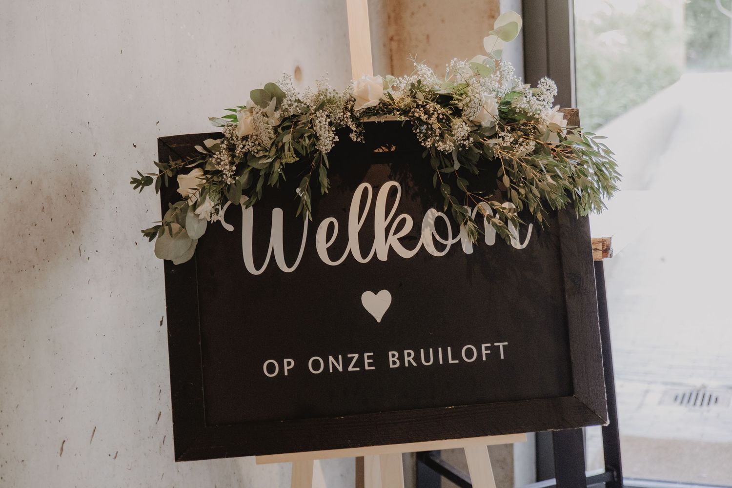 Elegant wedding welcome sign reading Welkom Op Onze Bruiloft decorated with white flowers and greenery on a dark chalkboard.