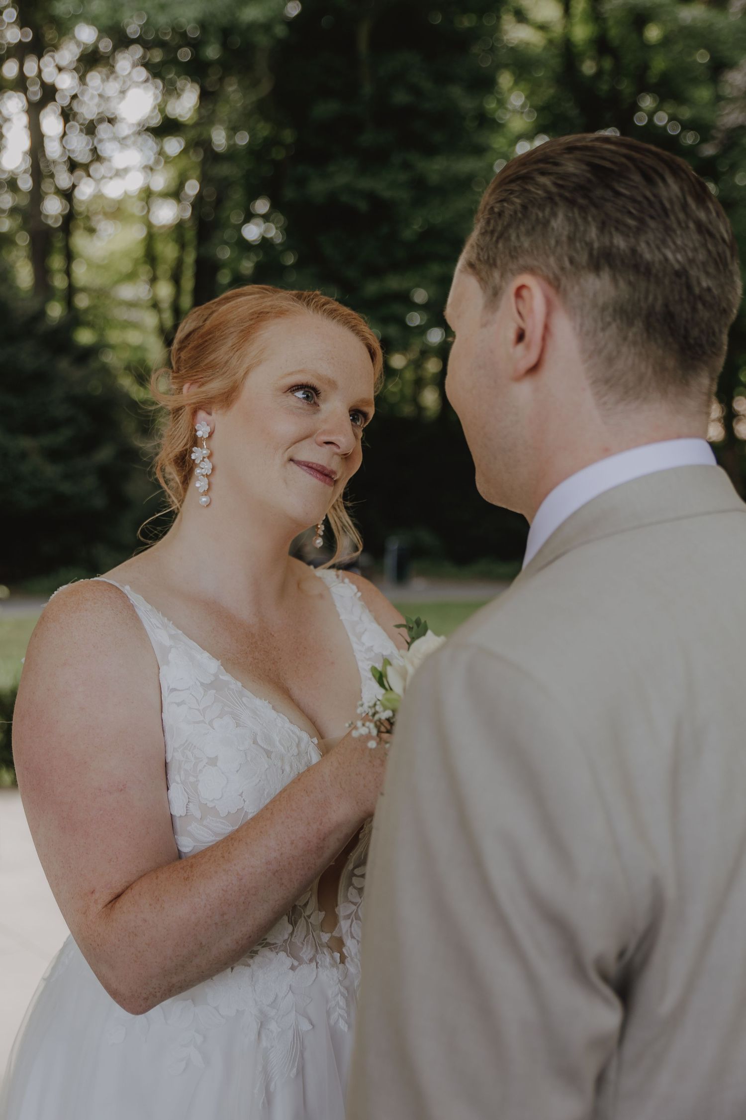 Radiant bride in lace gown gazes lovingly at groom during outdoor forest wedding ceremony.
