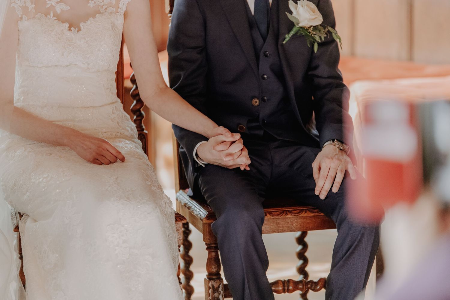 Bride and groom holding hands while seated during wedding ceremony, groom wearing dark suit with boutonniere.