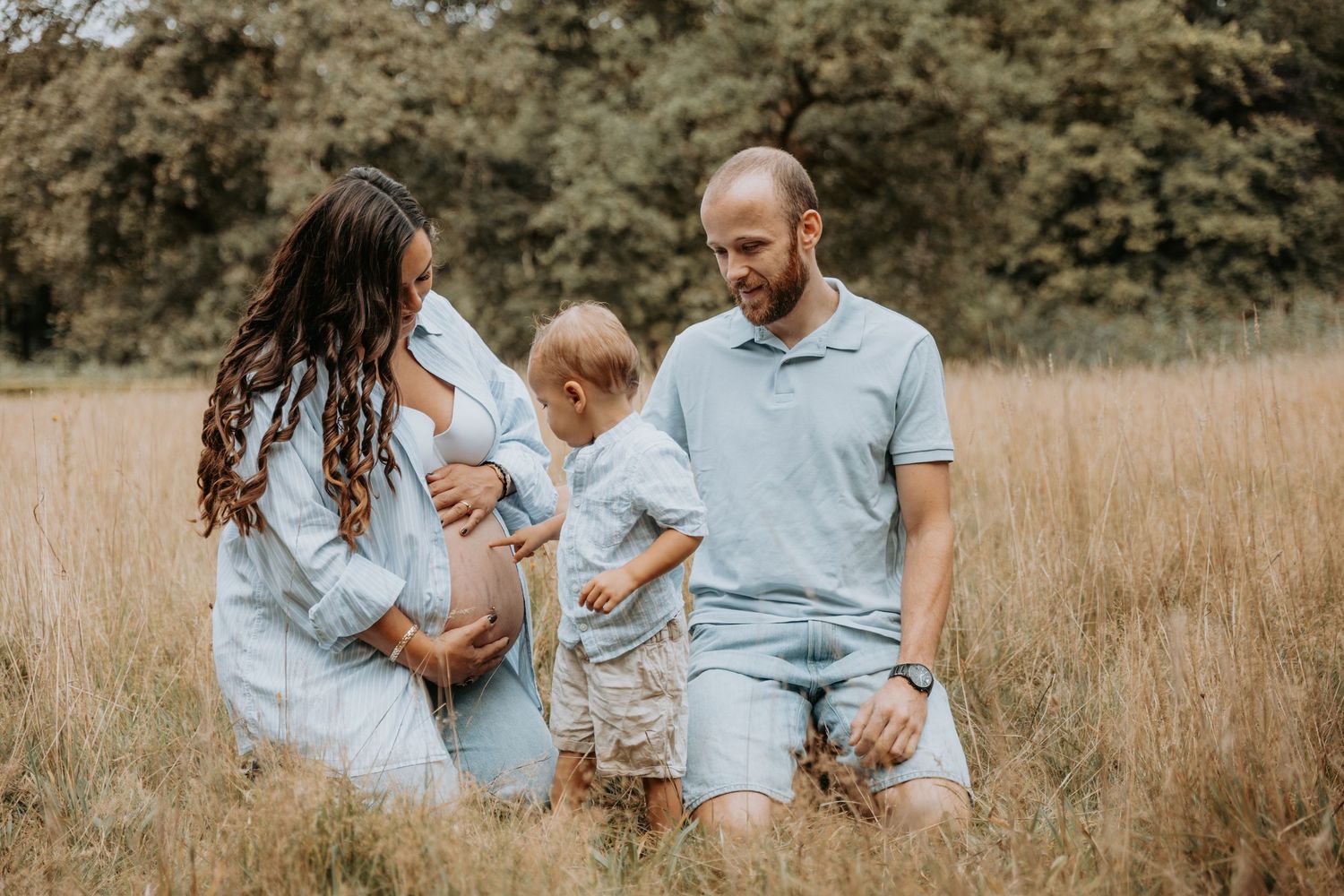Pregnant woman and man kneel in golden field with toddler son during outdoor maternity photo session.