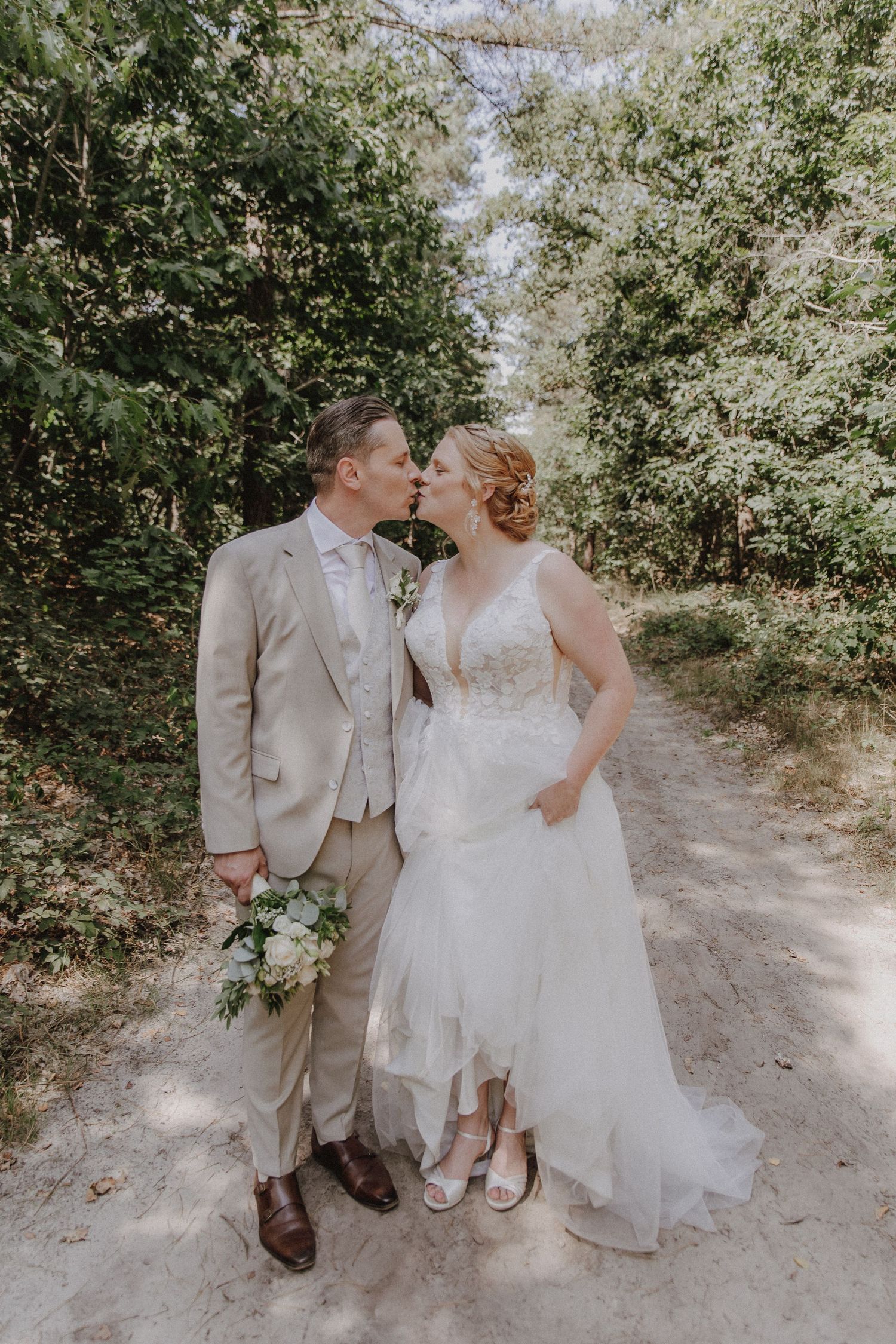 Bride and groom kissing on sandy forest path, bride in white gown, groom in beige suit.