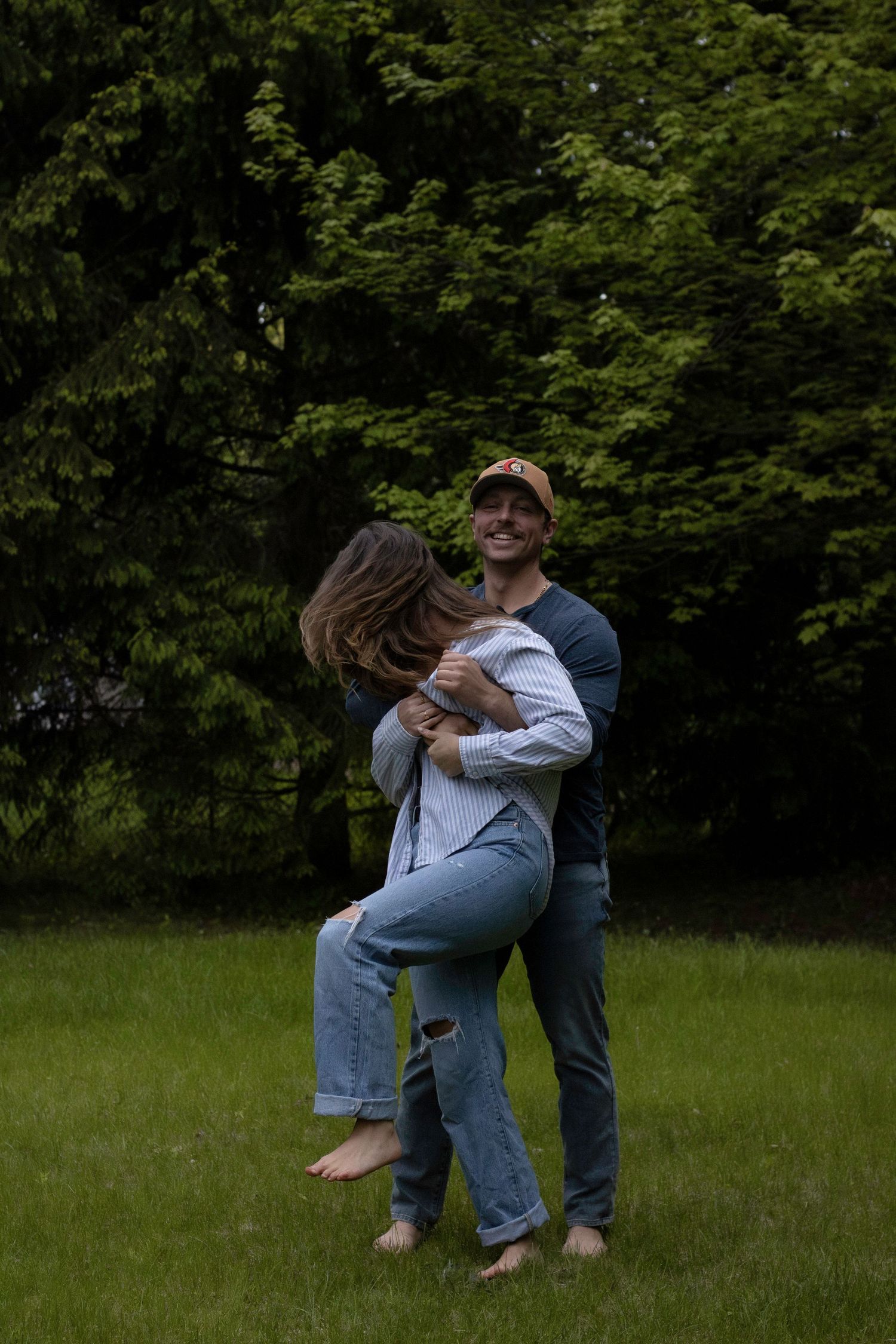 A couple embraces joyfully while standing barefoot in a grass field with lush green trees in the background.