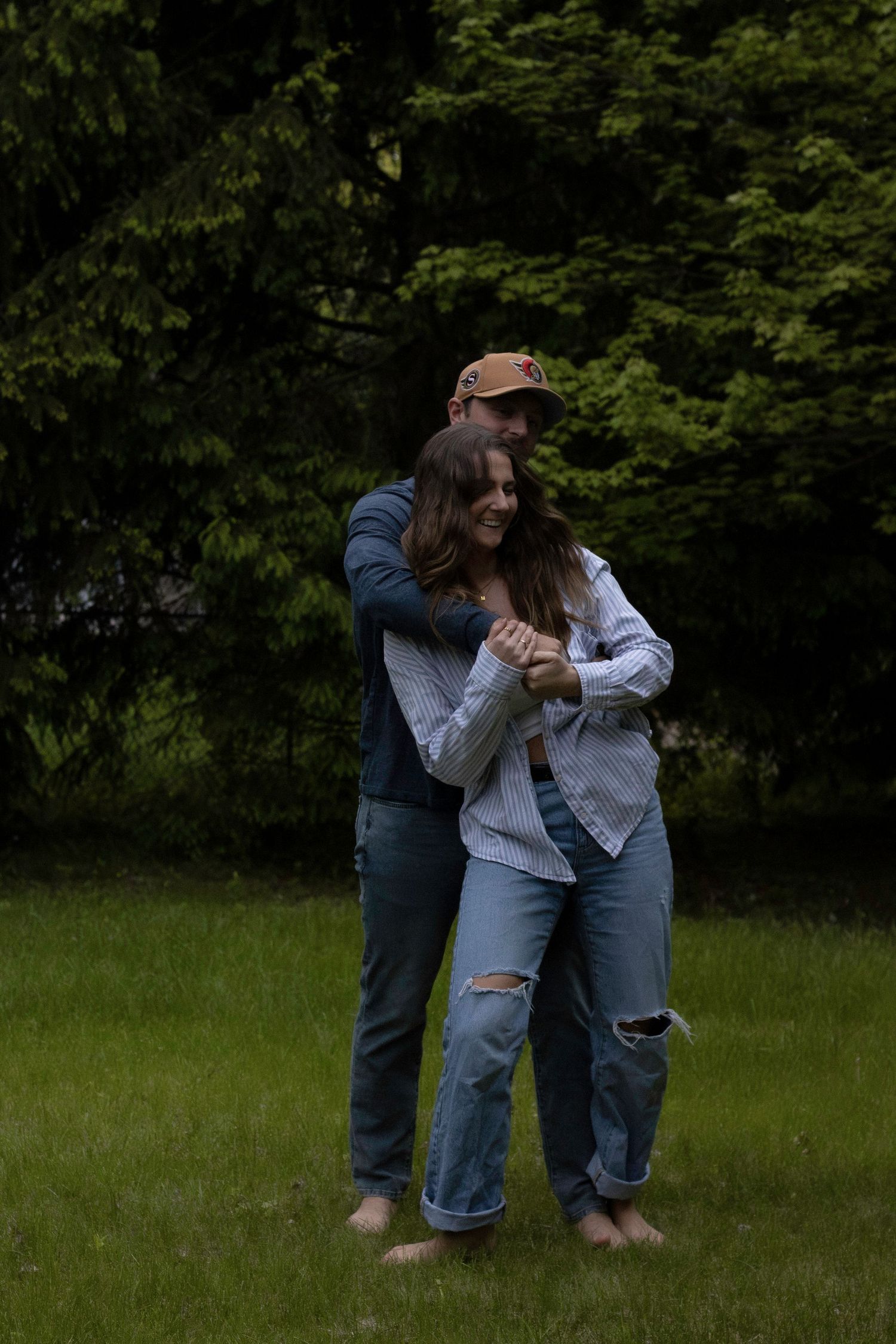 A couple in casual denim embraces while standing in a green grassy field with trees in the background.