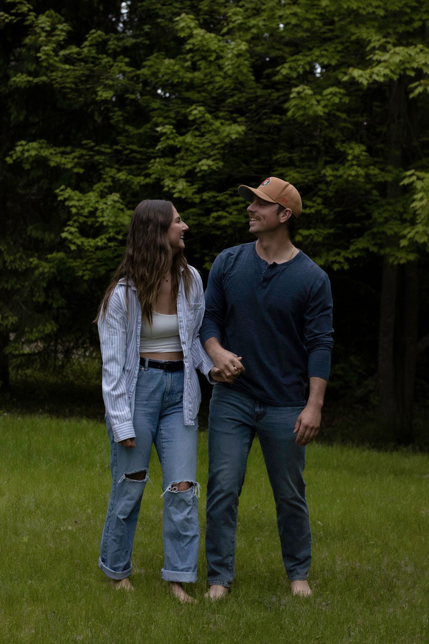 A couple in casual denim outfits standing together on a green lawn with forest backdrop.