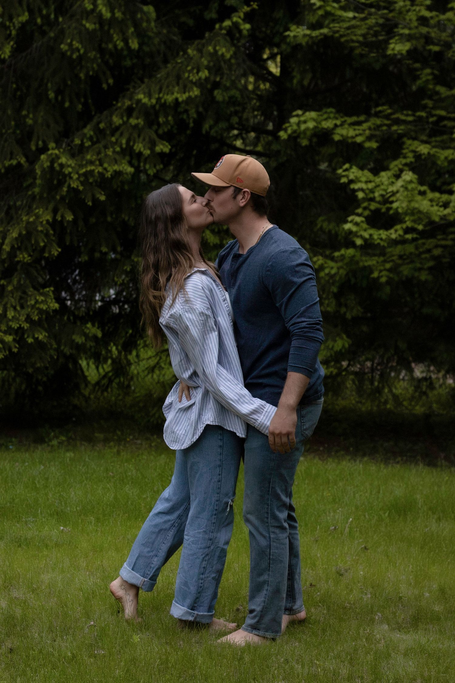 A romantic couple embraces barefoot in a grassy meadow with evergreen trees in the background.