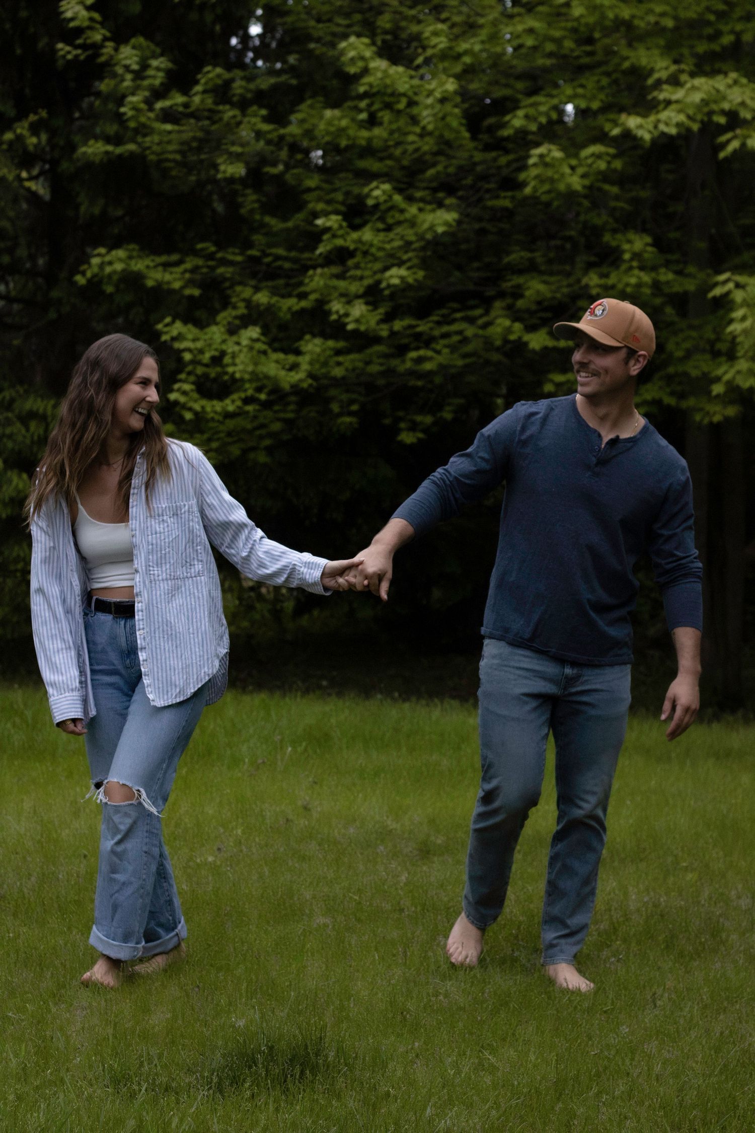 A couple holds hands while walking barefoot on grass with trees in background.