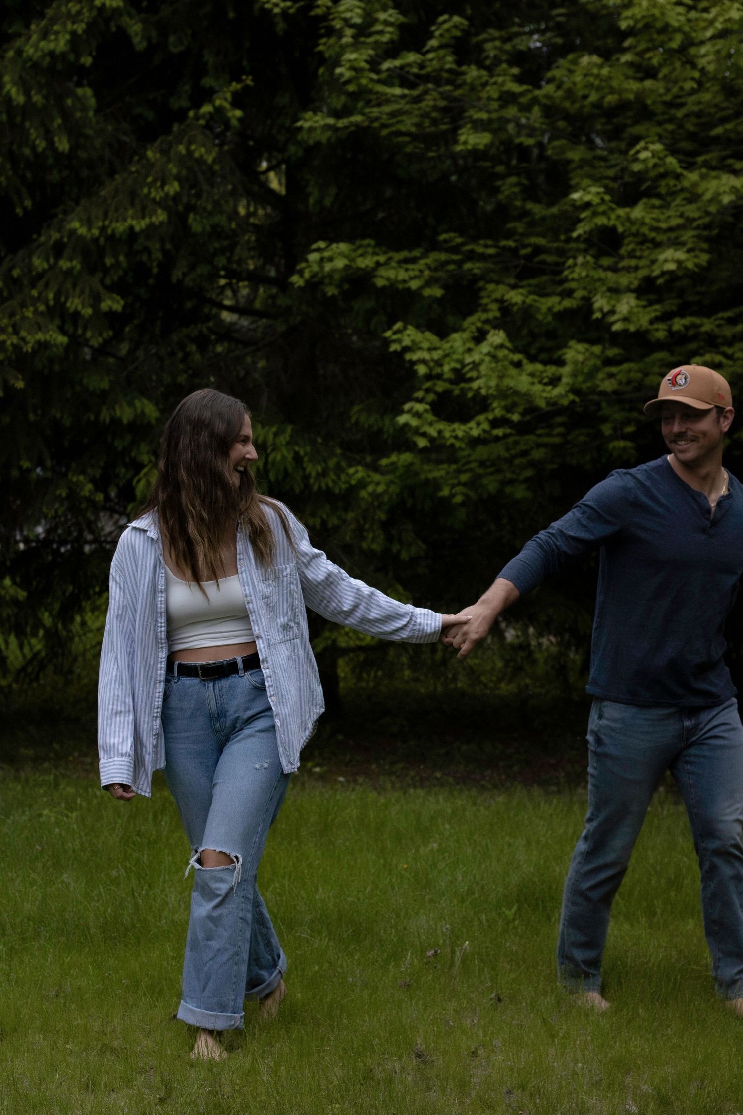 Two people holding hands in a grassy outdoor area surrounded by lush green trees on a cloudy day.