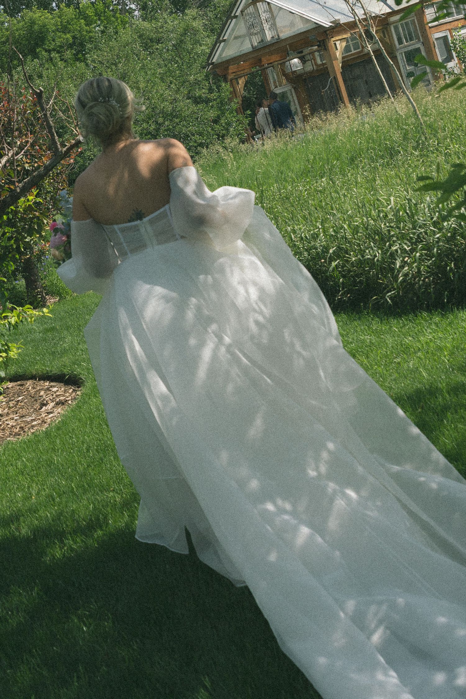 Flowing white wedding dress trails behind on green garden path.