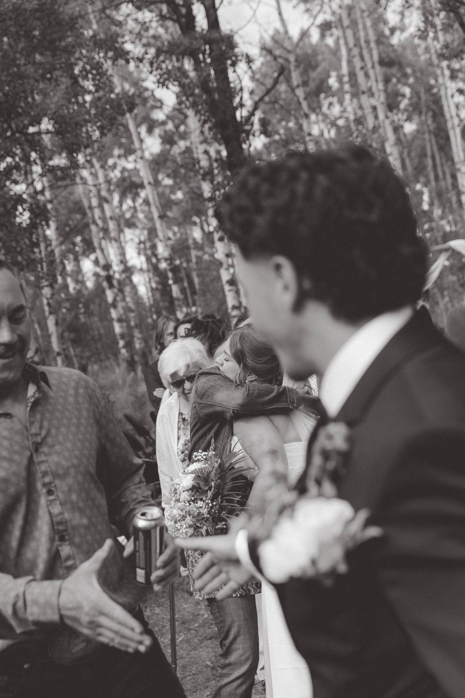 Black and white outdoor wedding scene with guests embracing among birch trees, groom in suit visible in foreground.