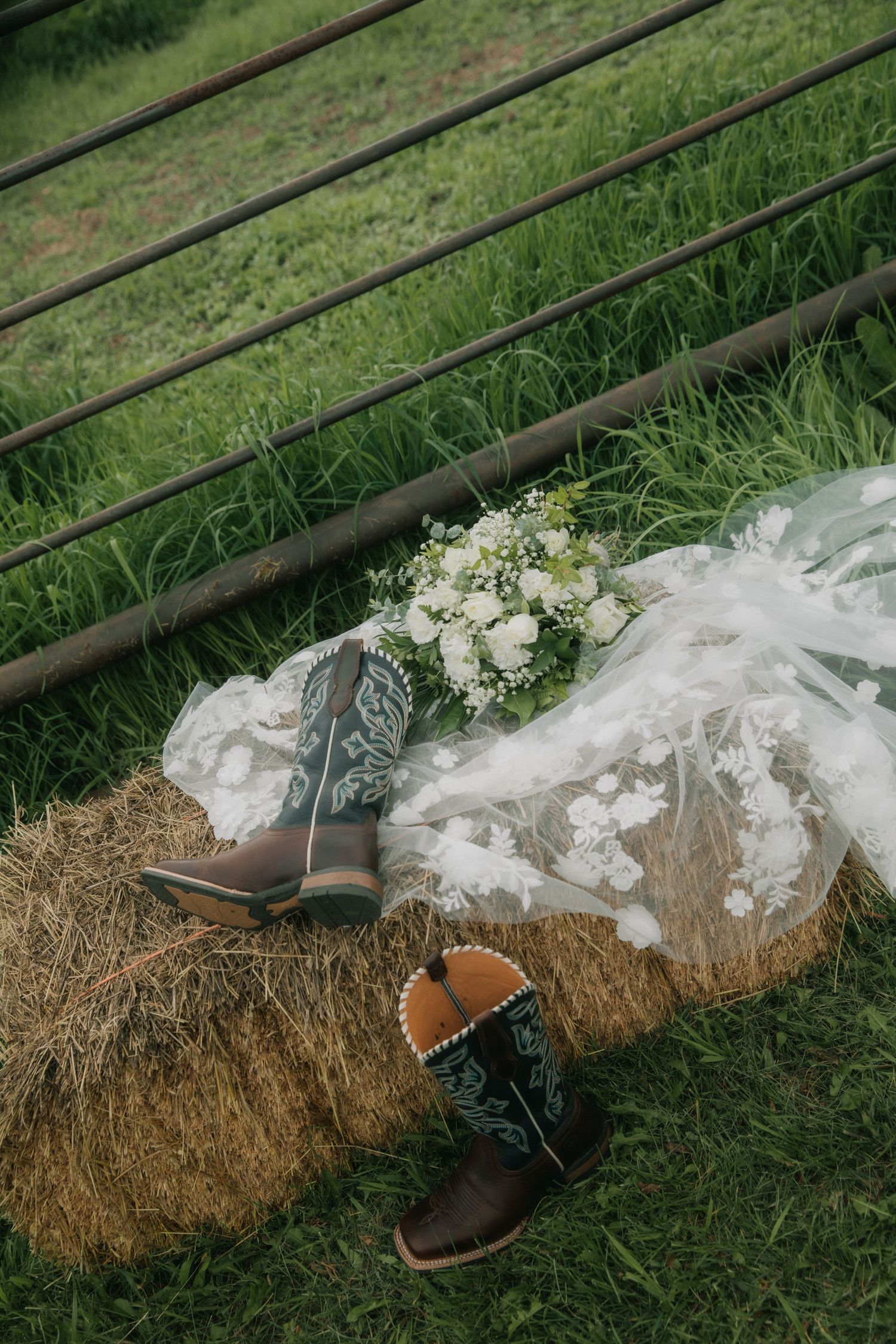 Cowboy boots and bridal veil with white bouquet resting on hay bale at farm wedding.