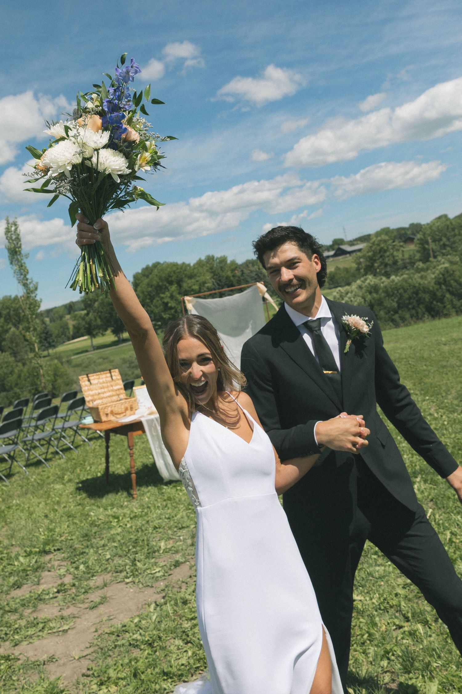 Joyful newlyweds celebrate after outdoor wedding ceremony, bride raises floral bouquet triumphantly as groom smiles in black suit.