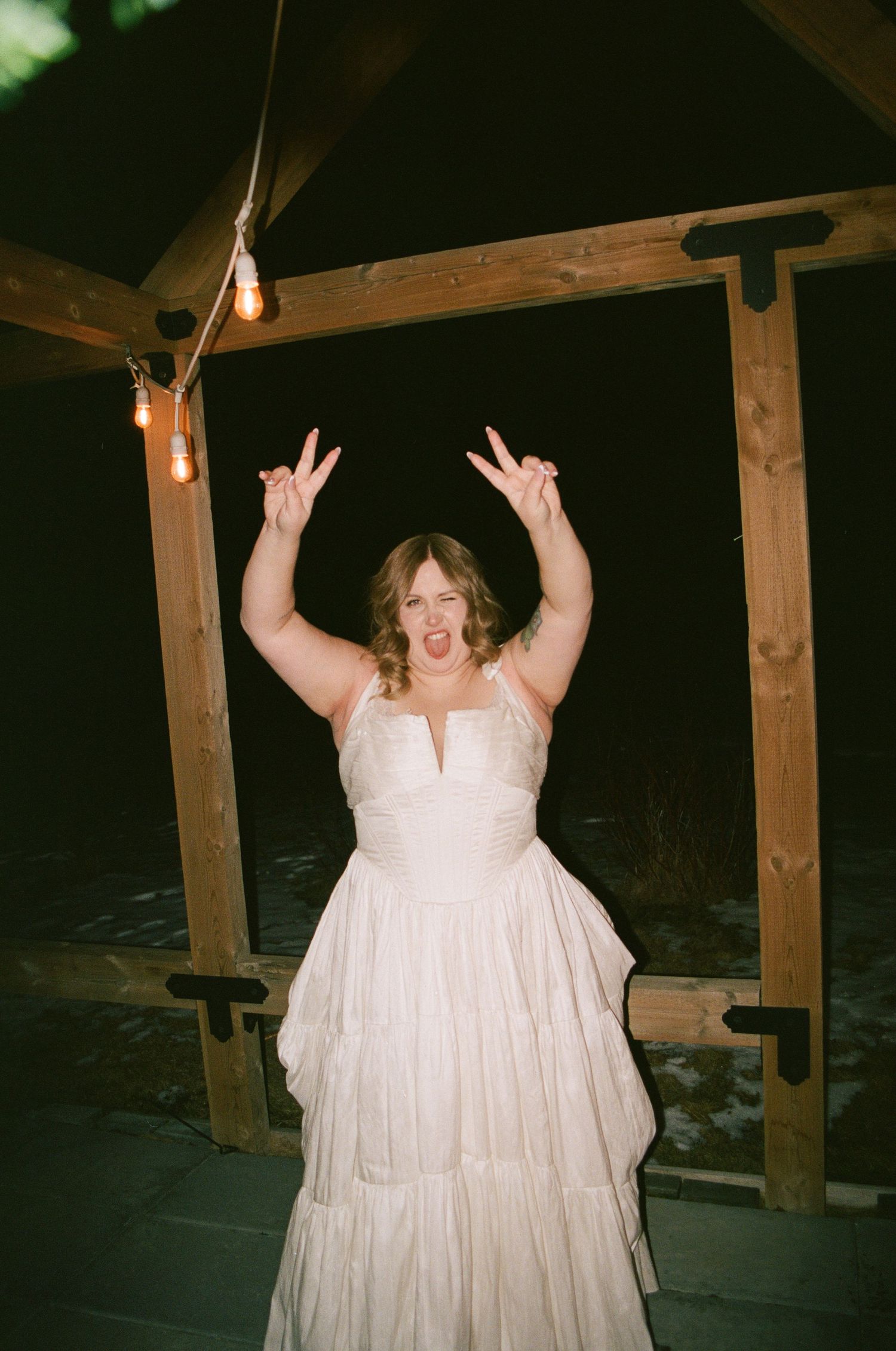 A joyful bride in a white ruffled gown raises her hands making rock signs under a wooden gazebo with string lights.