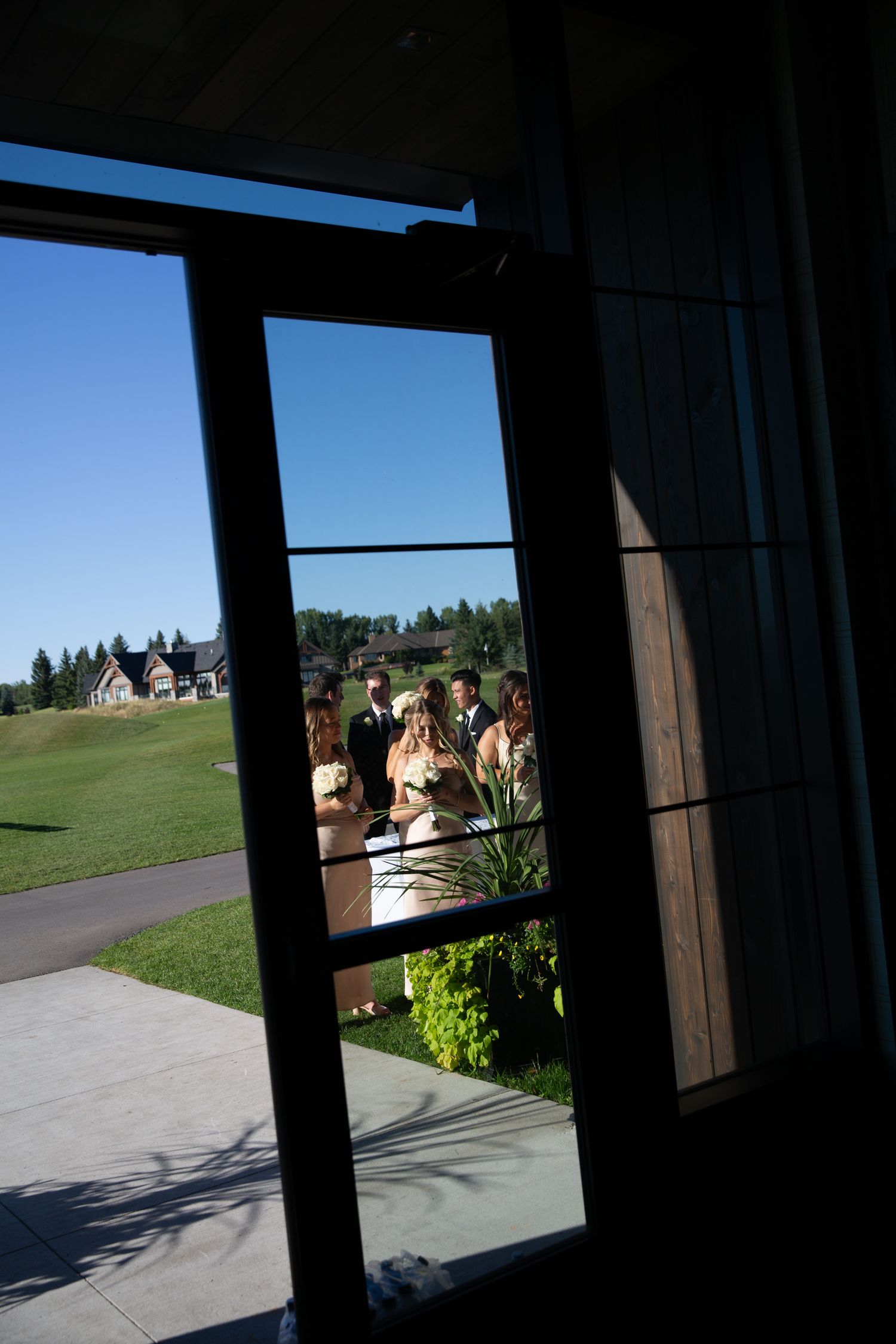 Wedding party visible through a window frame against a landscaped outdoor backdrop.