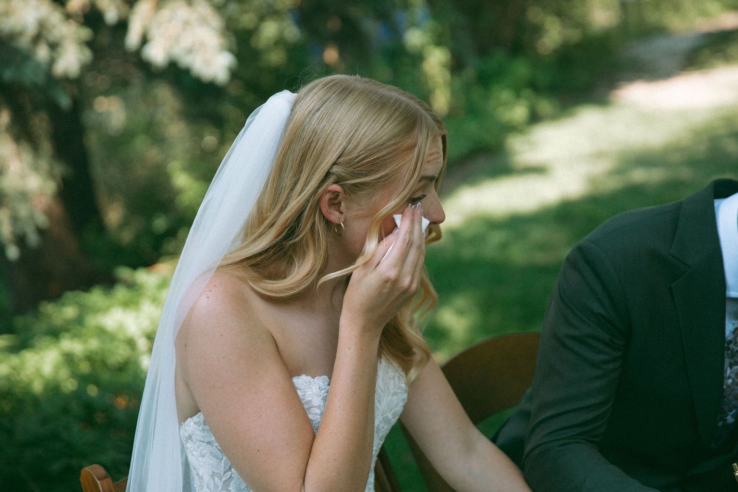 An emotional bride in a white strapless wedding dress wipes tears during an outdoor ceremony in a garden setting.