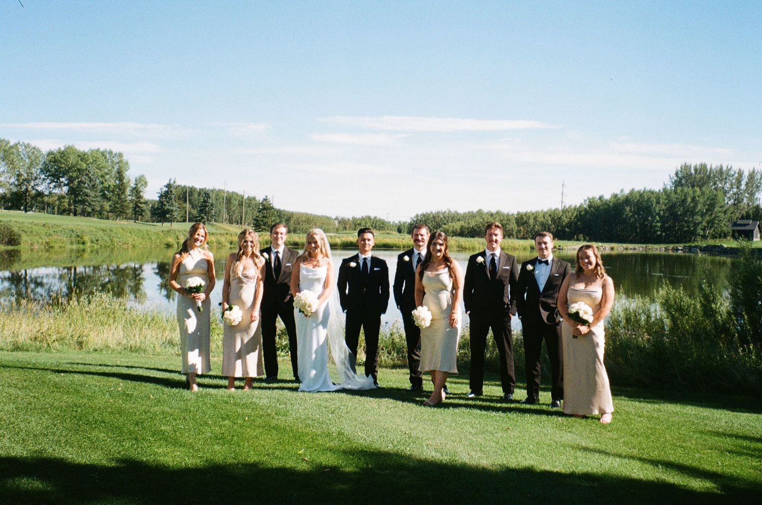 Wedding party stands together on green lawn near scenic pond and treeline on sunny summer day.