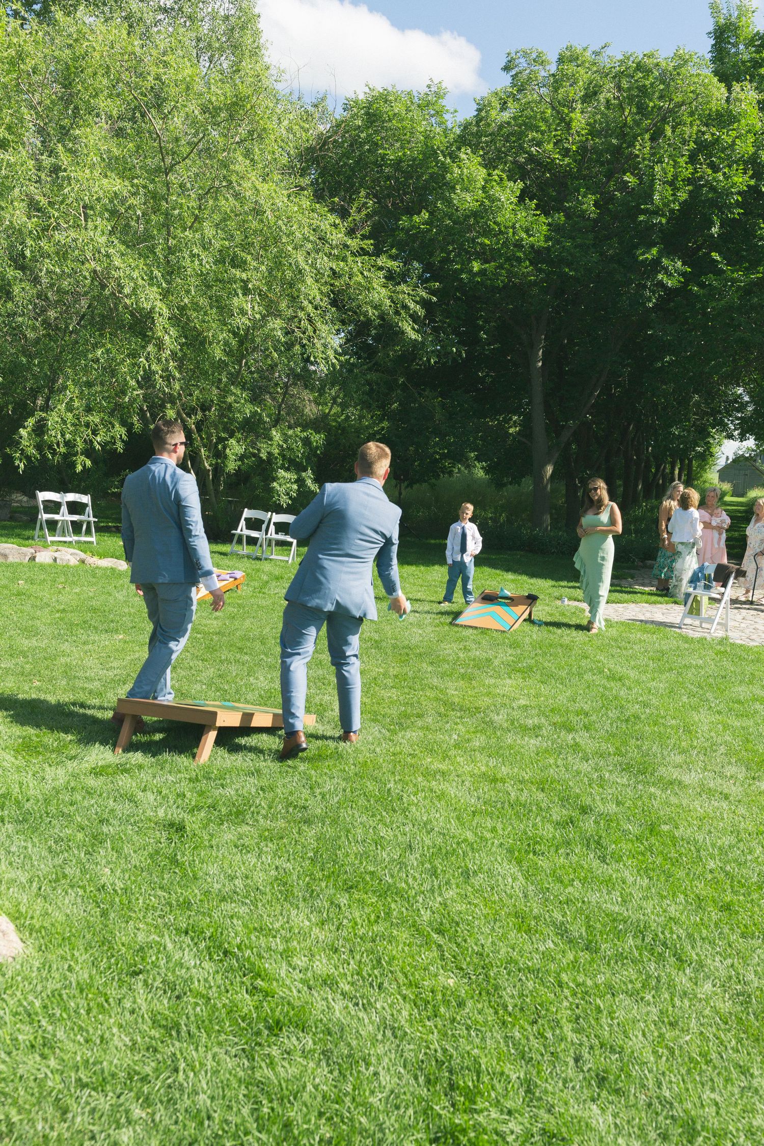 Wedding guests mingle on a sunny green lawn with trees in the background.