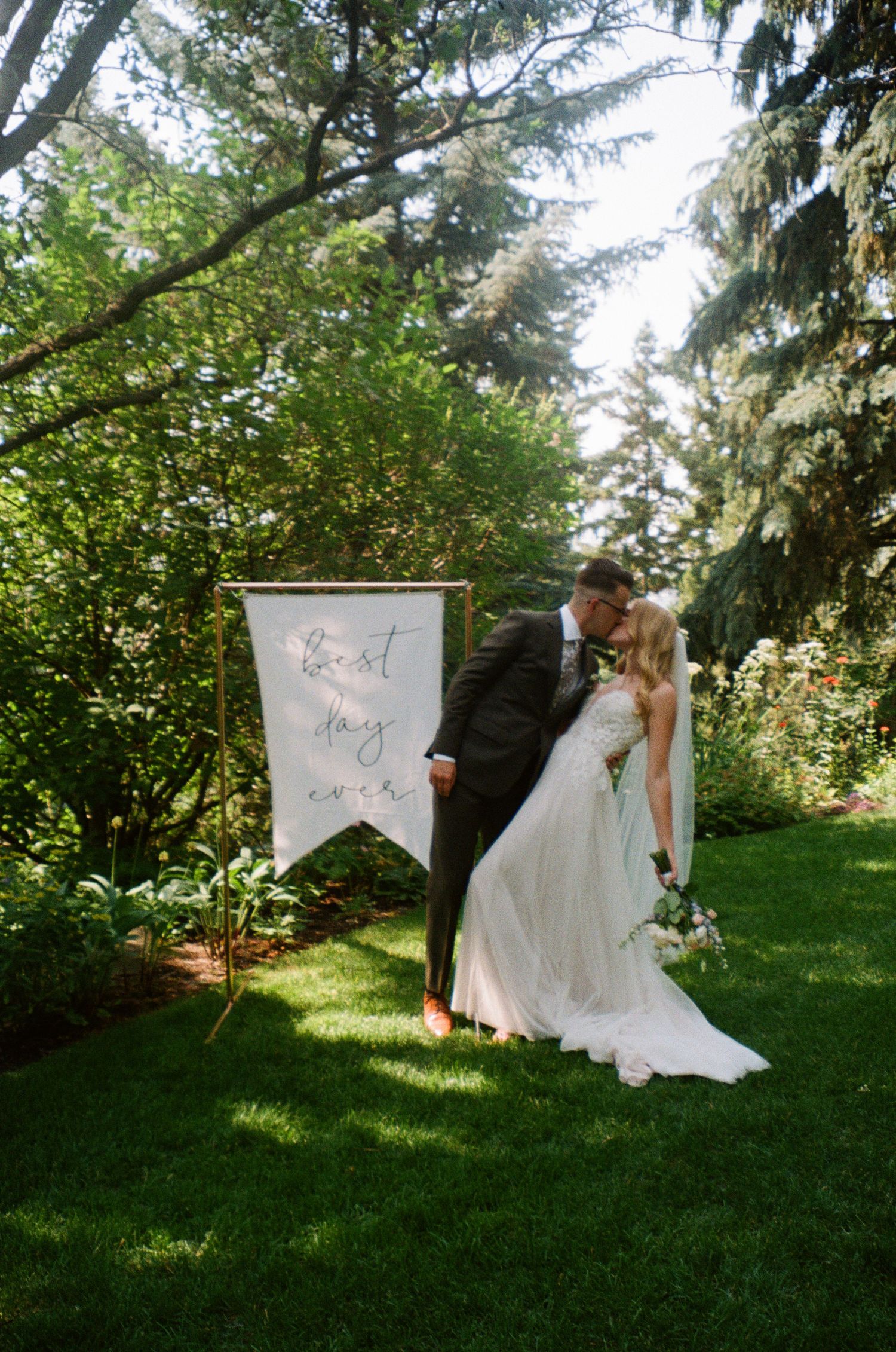 A couple shares a romantic moment in a sunlit garden setting with a decorative white banner and lush greenery.