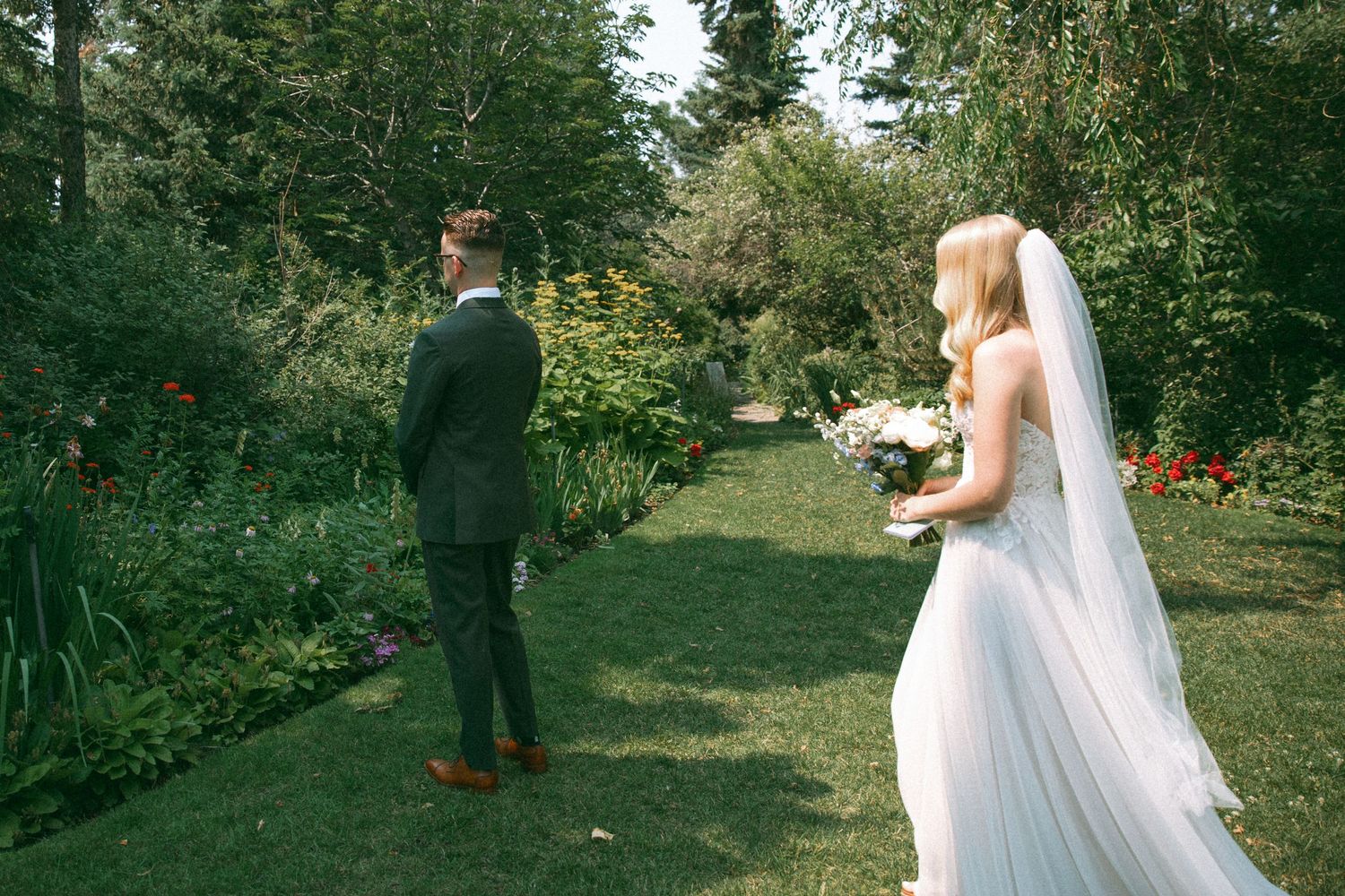A wedding couple stands in a lush garden setting with blooming flowers and evergreen trees.