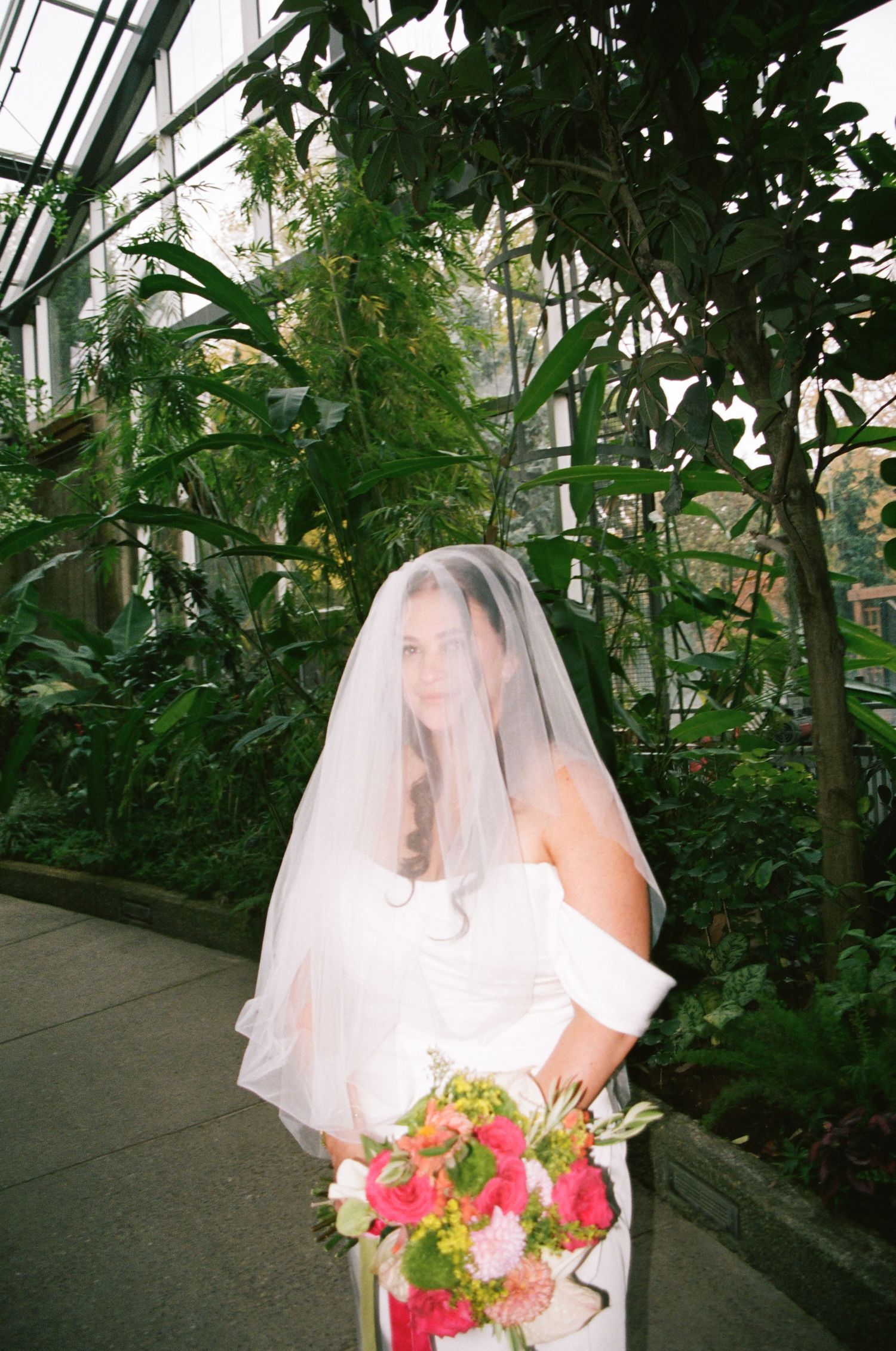 Bride in white strapless gown and veil holds pink bouquet inside lush green botanical garden greenhouse.