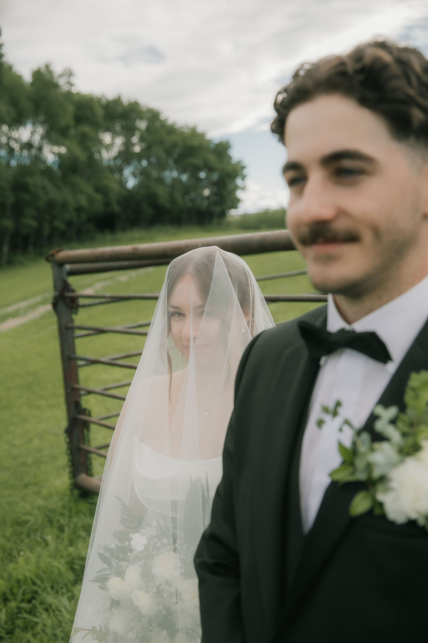 Bride in veil standing behind groom in black tuxedo at rustic outdoor farm wedding.