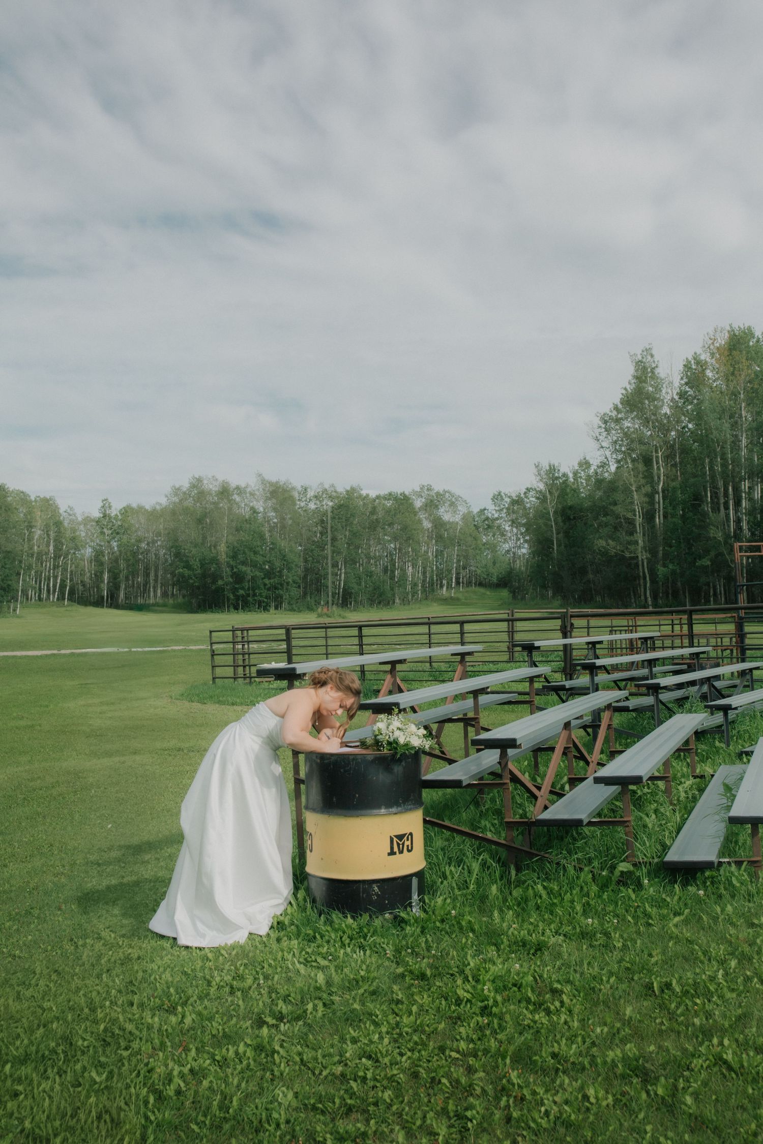 Bride in white gown placing flowers in a barrel planter among picnic tables at an outdoor wedding venue.