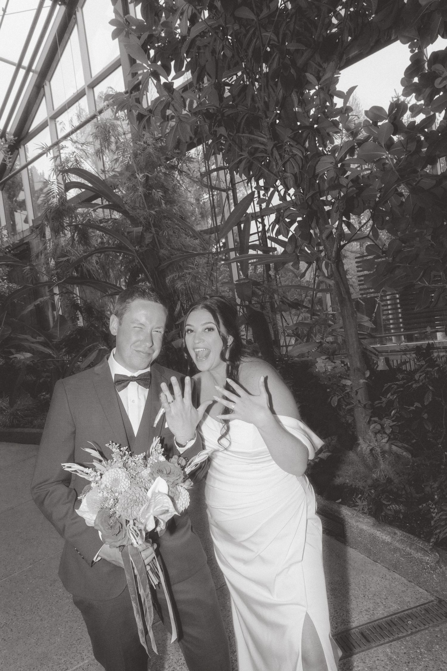 Black and white photo of newlyweds showing wedding rings, bride in white gown holding bouquet in a lush greenhouse venue.