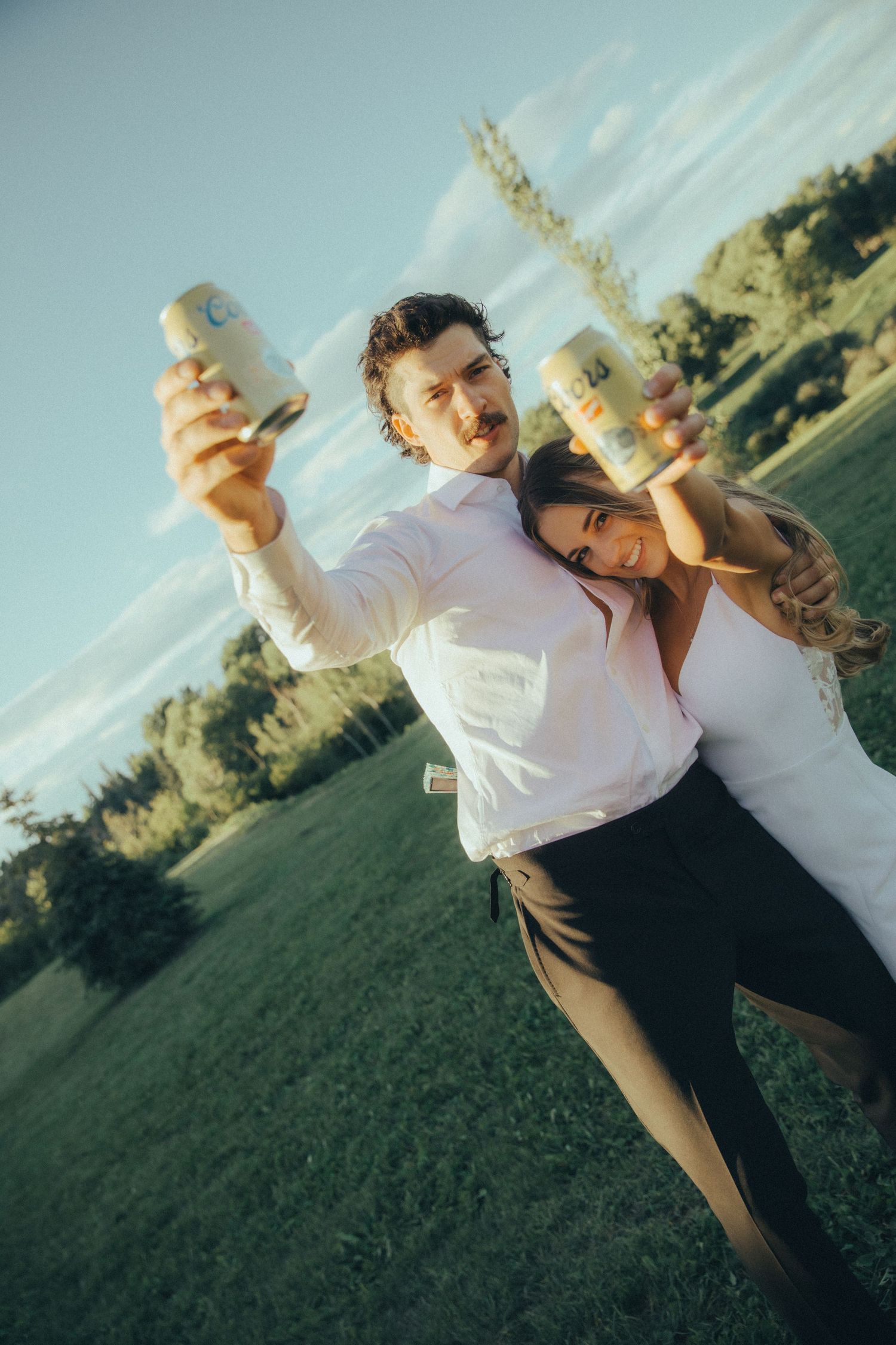 Joyful newlyweds toast with beer cans outdoors on a sunny green field after their wedding ceremony.