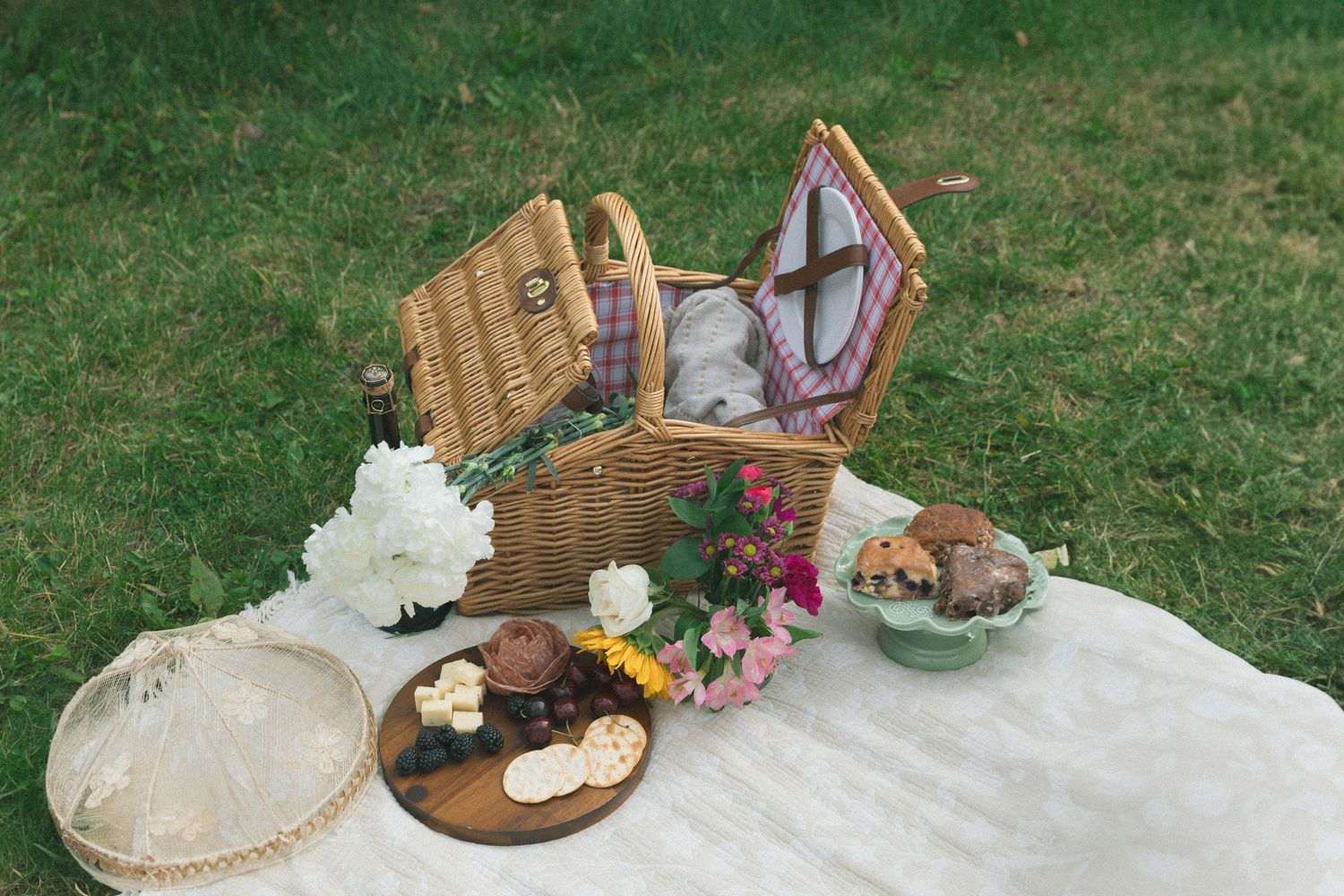 Vintage picnic basket with wine glasses, baked goods, and flowers arranged on white blanket in green grass.