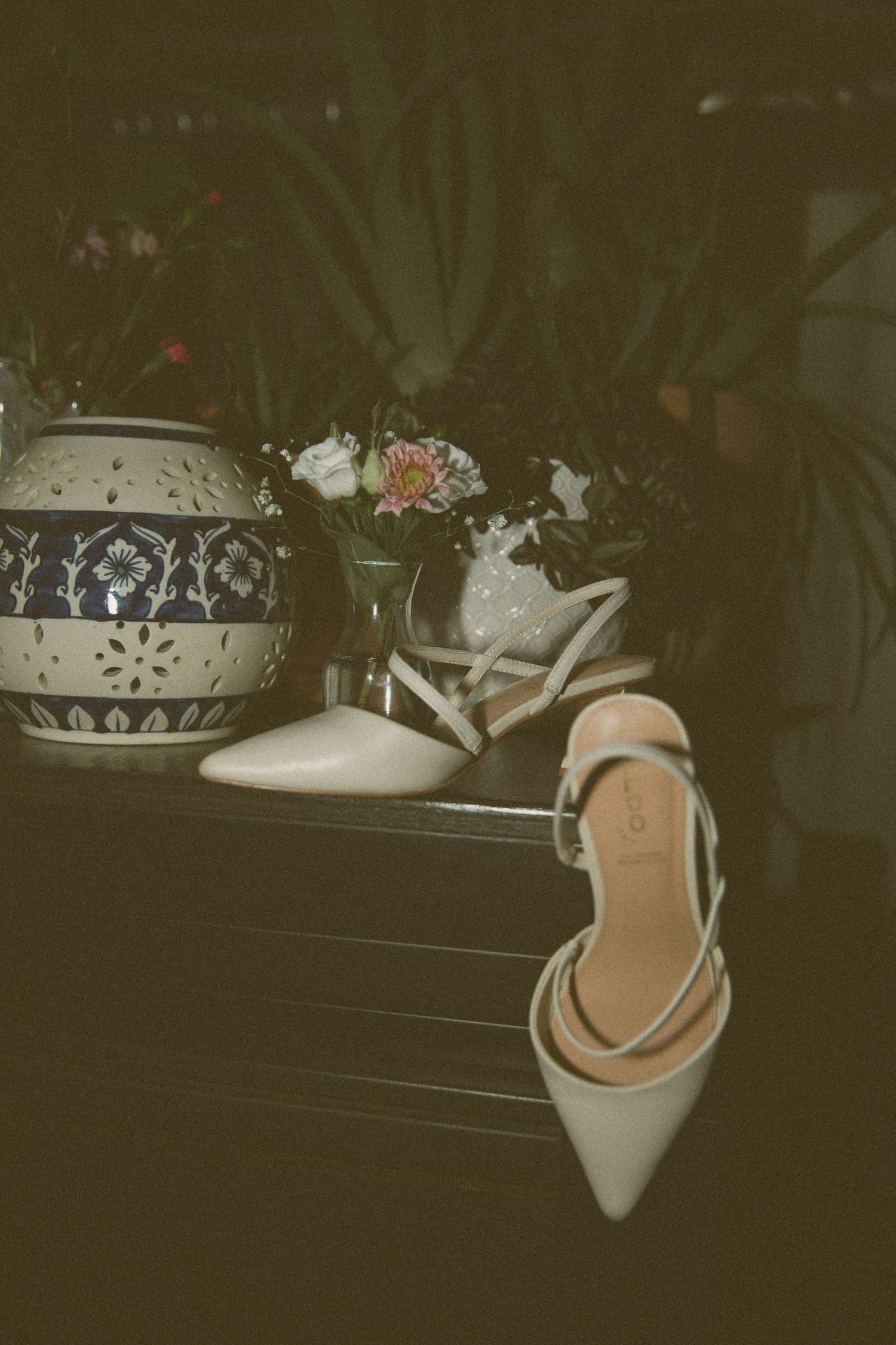 A vintage-style photo showing an elegant beige slingback heel next to decorative vases with flowers in dark lighting.