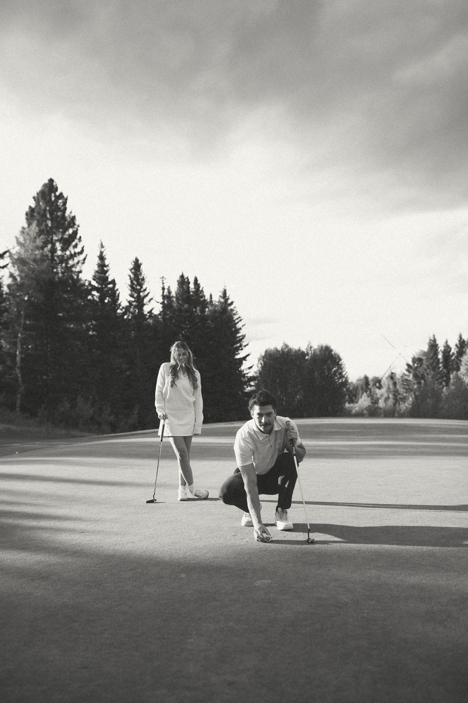 Two golfers line up their putts on a scenic golf course surrounded by evergreen trees in black and white.