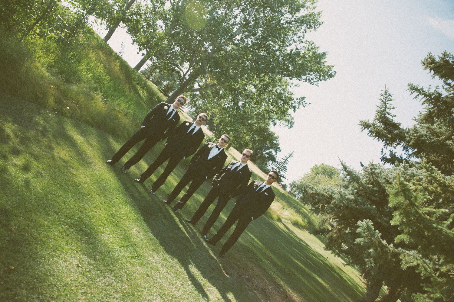 Wedding party members stand in a row outdoors on a grassy hillside under trees.