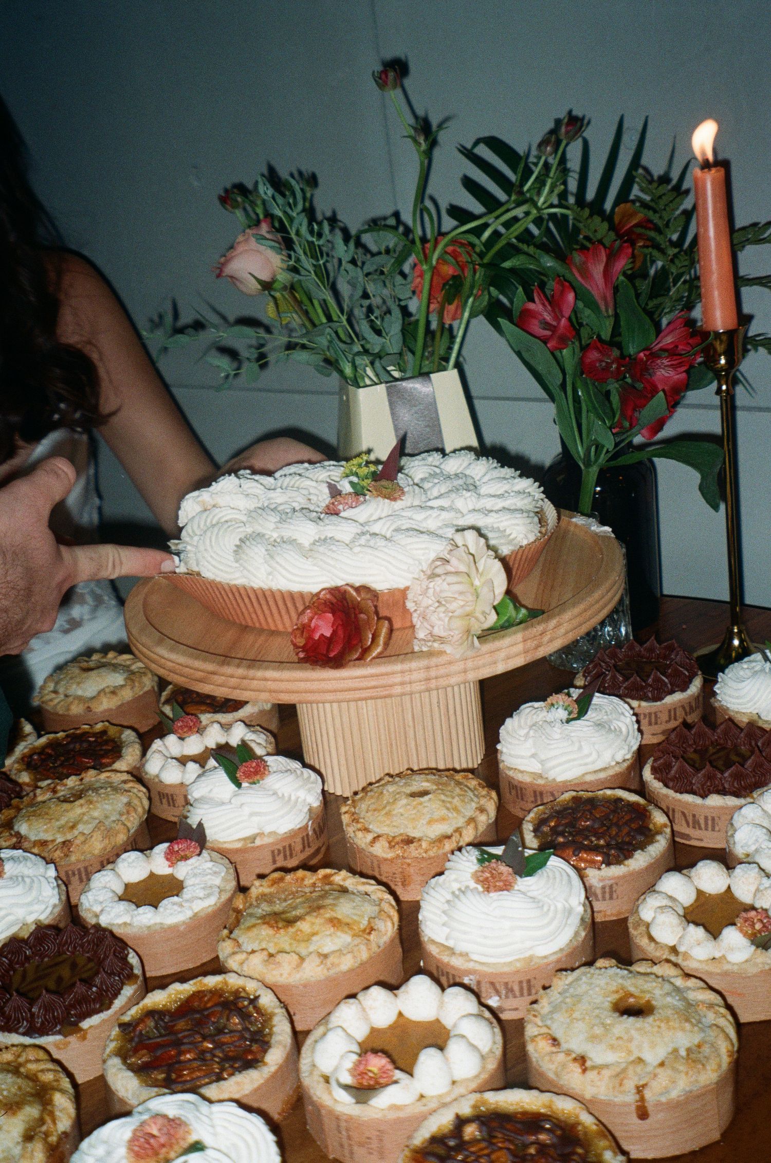 A vintage party scene featuring assorted cakes and desserts on wooden platters with flowers and a lit candle in the background.