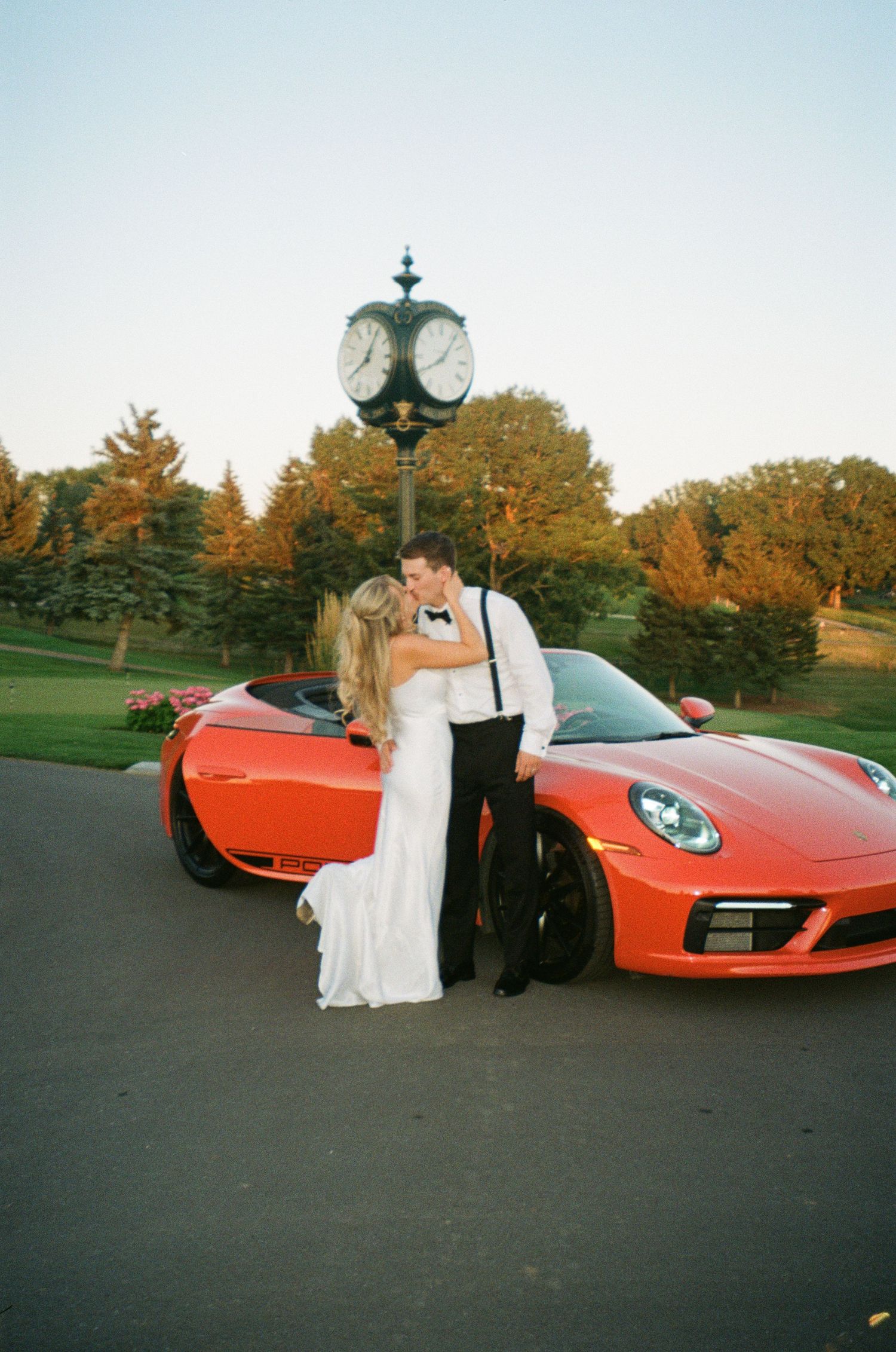 Bride and groom kissing beside a red Porsche 911 with a clock tower and autumn trees at golden hour.