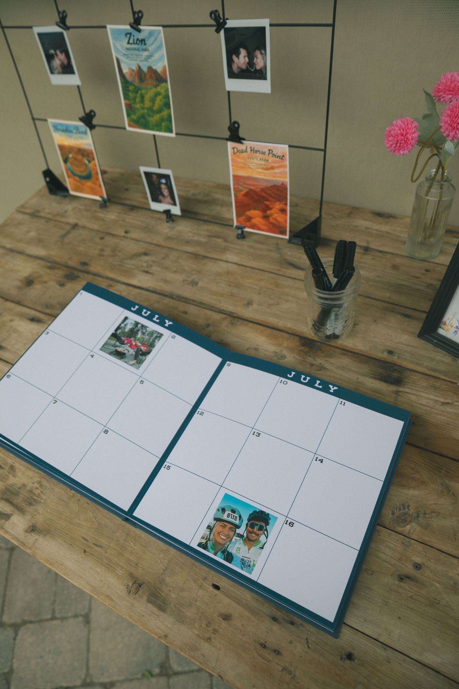 Calendar planner with photos displayed on wooden desk alongside pink flowers in a vase.